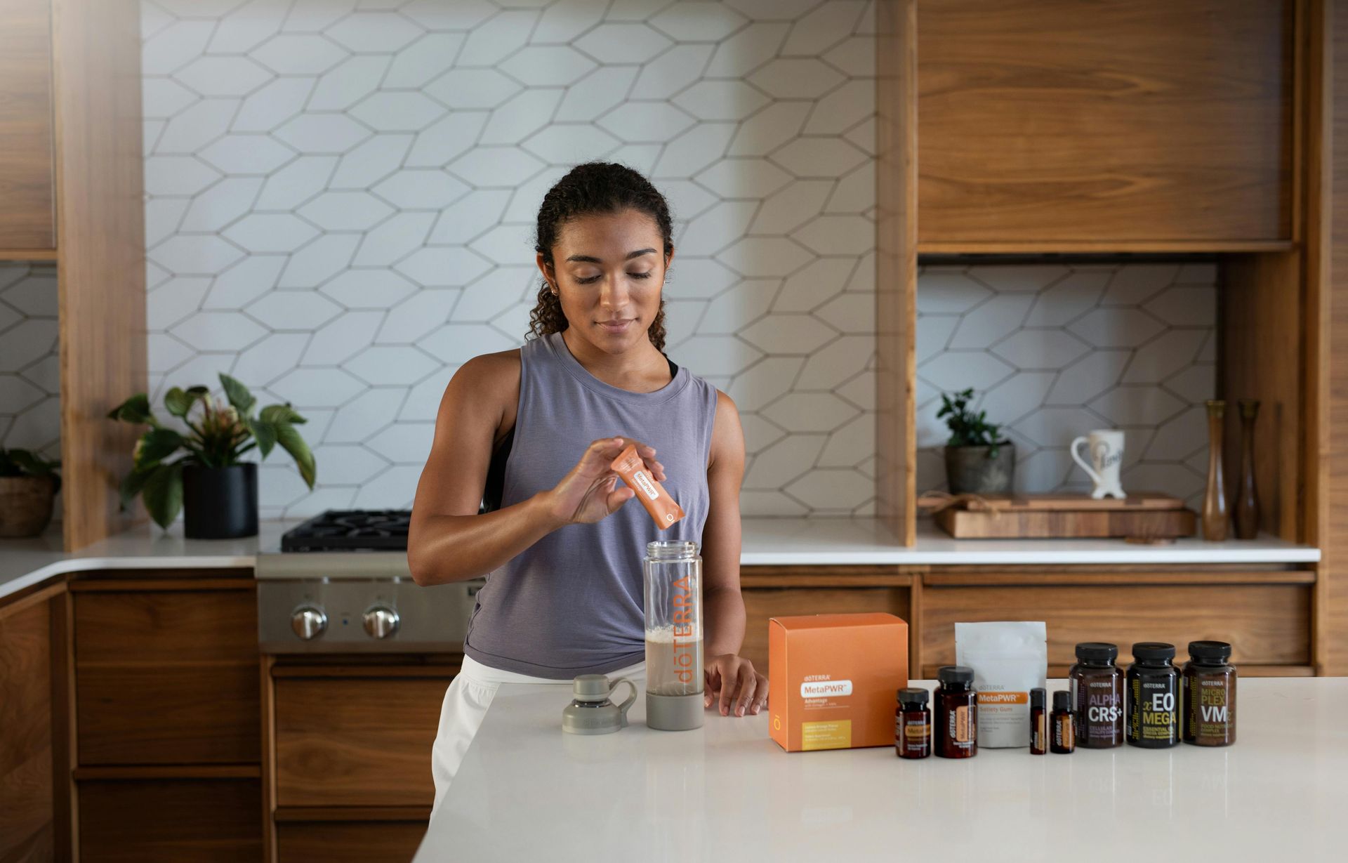 Woman mixing a drink in a kitchen beside assorted supplement bottles and an orange box.