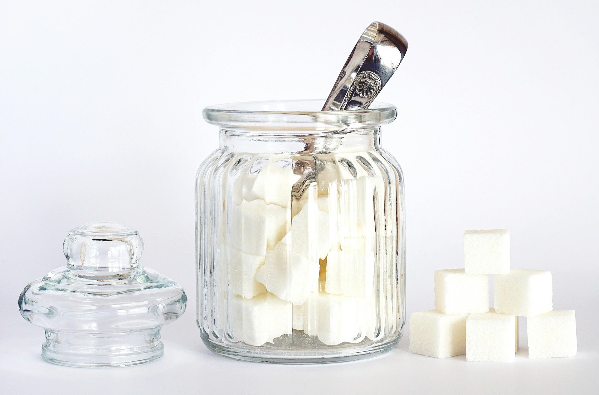 Glass jar of sugar cubes with metal tongs, alongside a sugar bowl and loose cubes on a white background