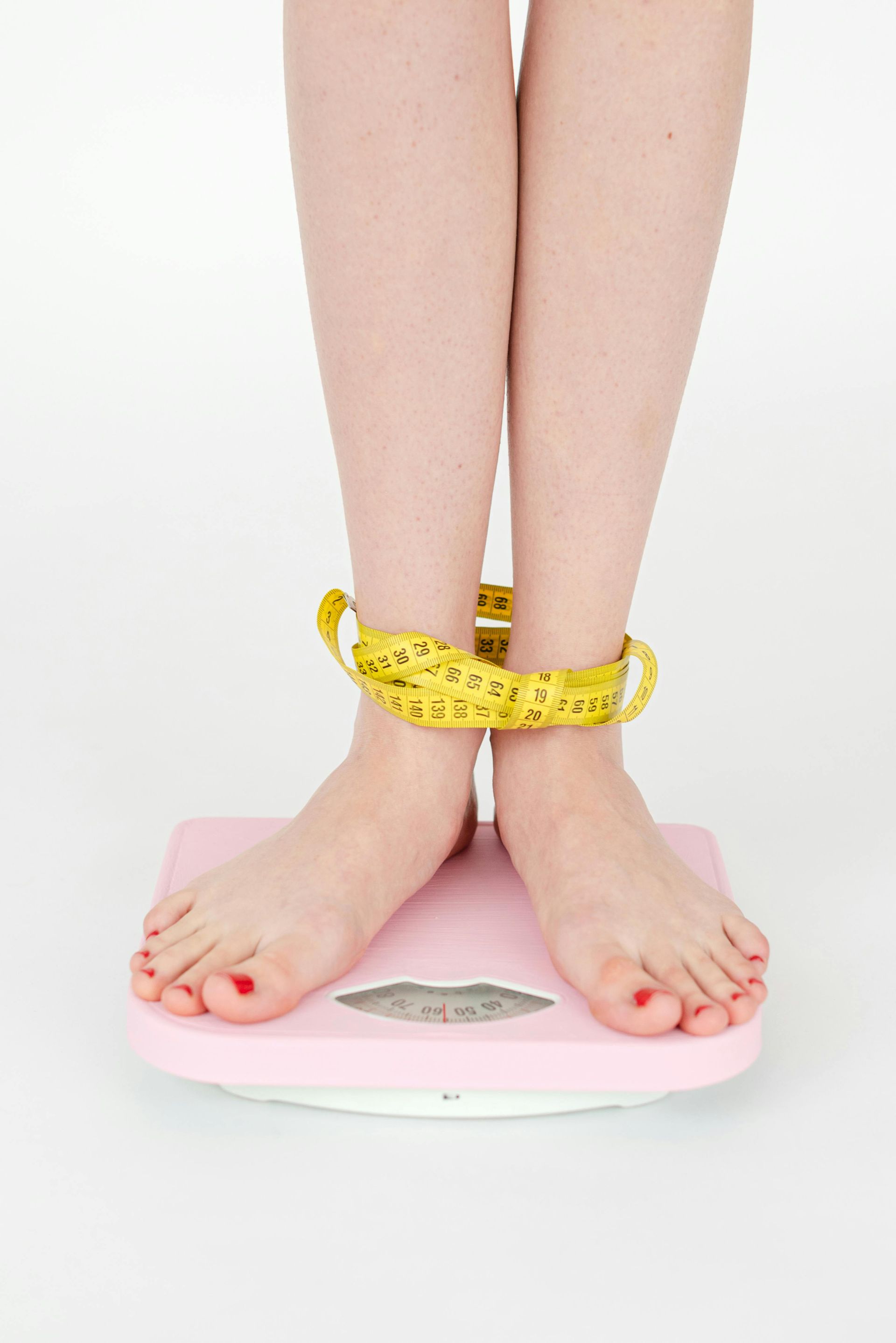 Feet standing on a pink scale with a yellow measuring tape wrapped around the ankles.