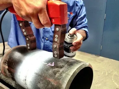 A person using a magnetic particle testing tool on a metal pipe, spraying it with a can of liquid.