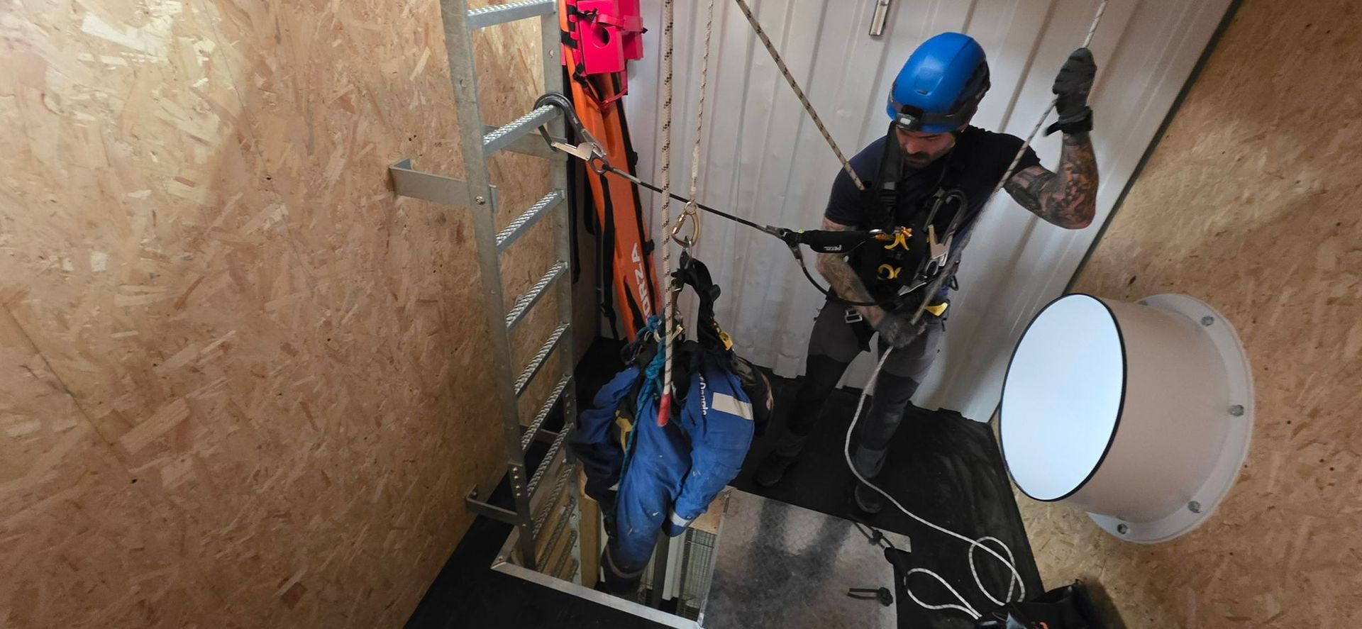 Worker in a blue helmet rappelling down a narrow industrial shaft with ropes and safety gear.