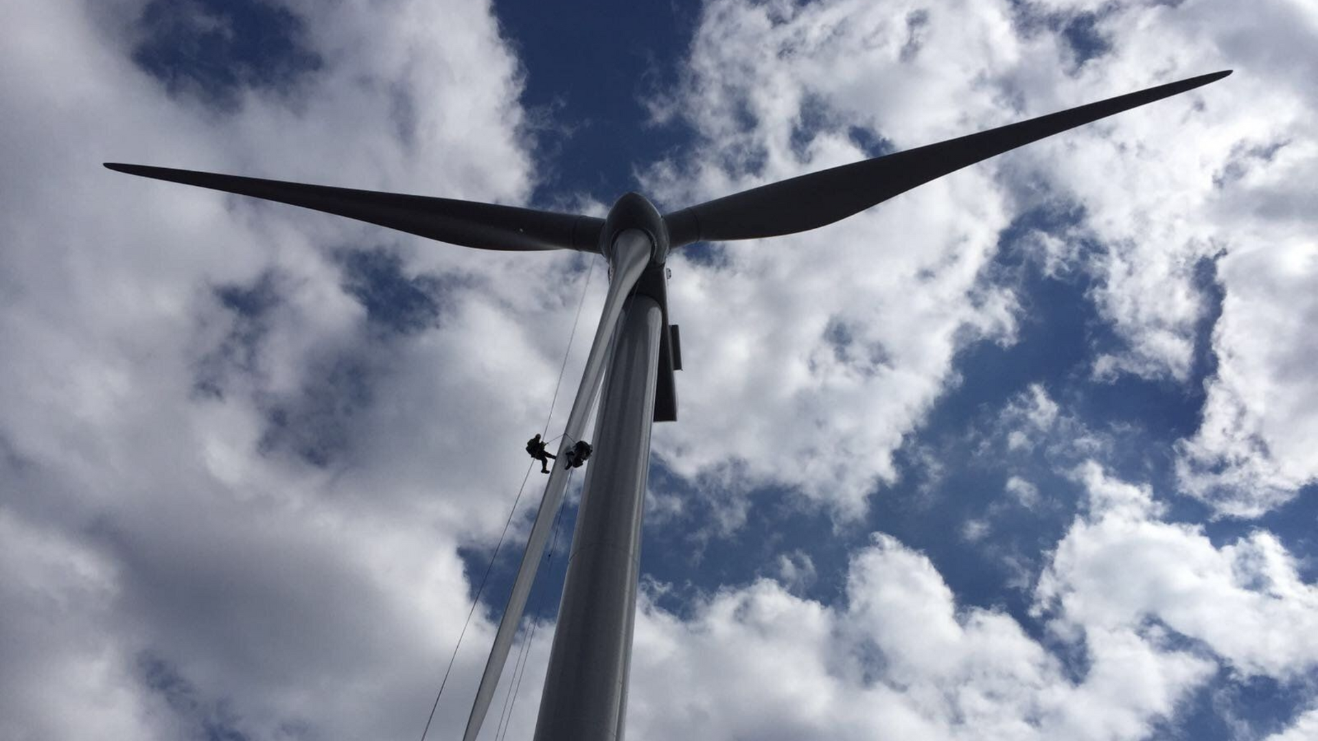 Wind turbine against a cloudy blue sky. Two abseilers cleaning a wind turbine blade.