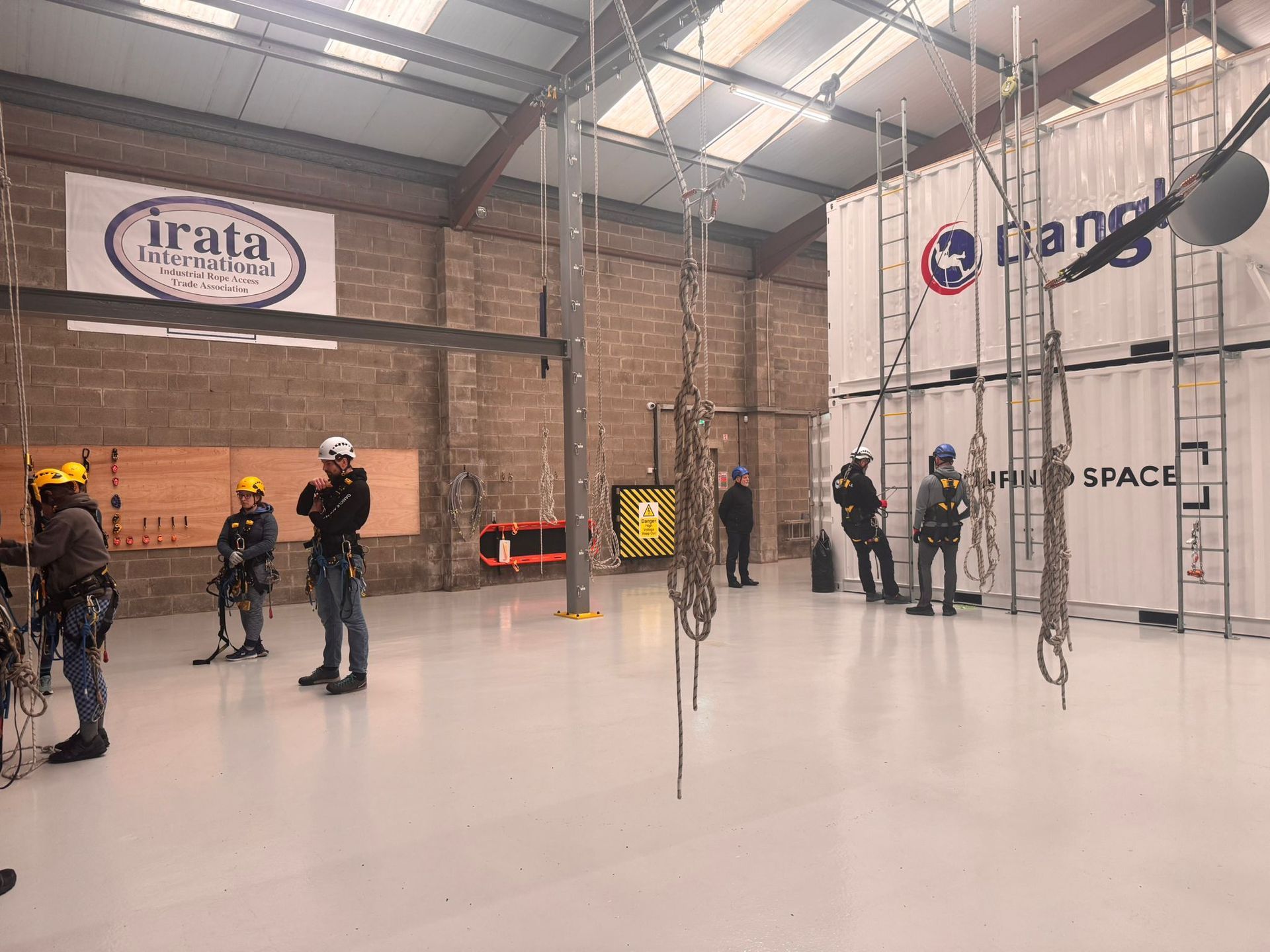 Several people in helmets stand in Dangle Academy with climbing ropes and hanging obstacles.