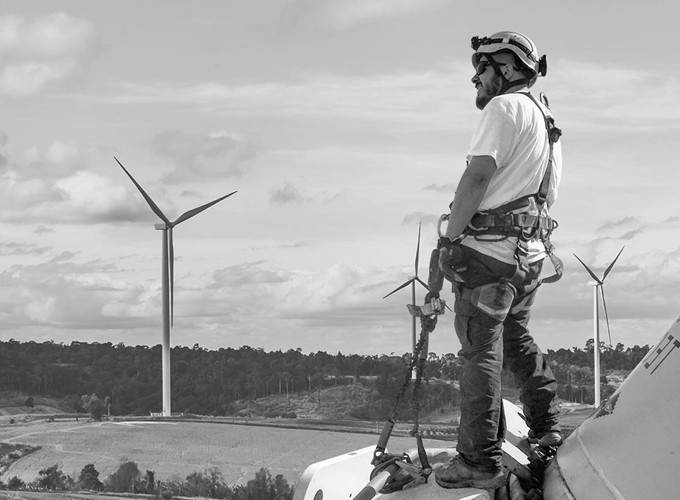 A wind turbine technician in safety gear stands on a turbine Nacelle overlooking a wind farm.