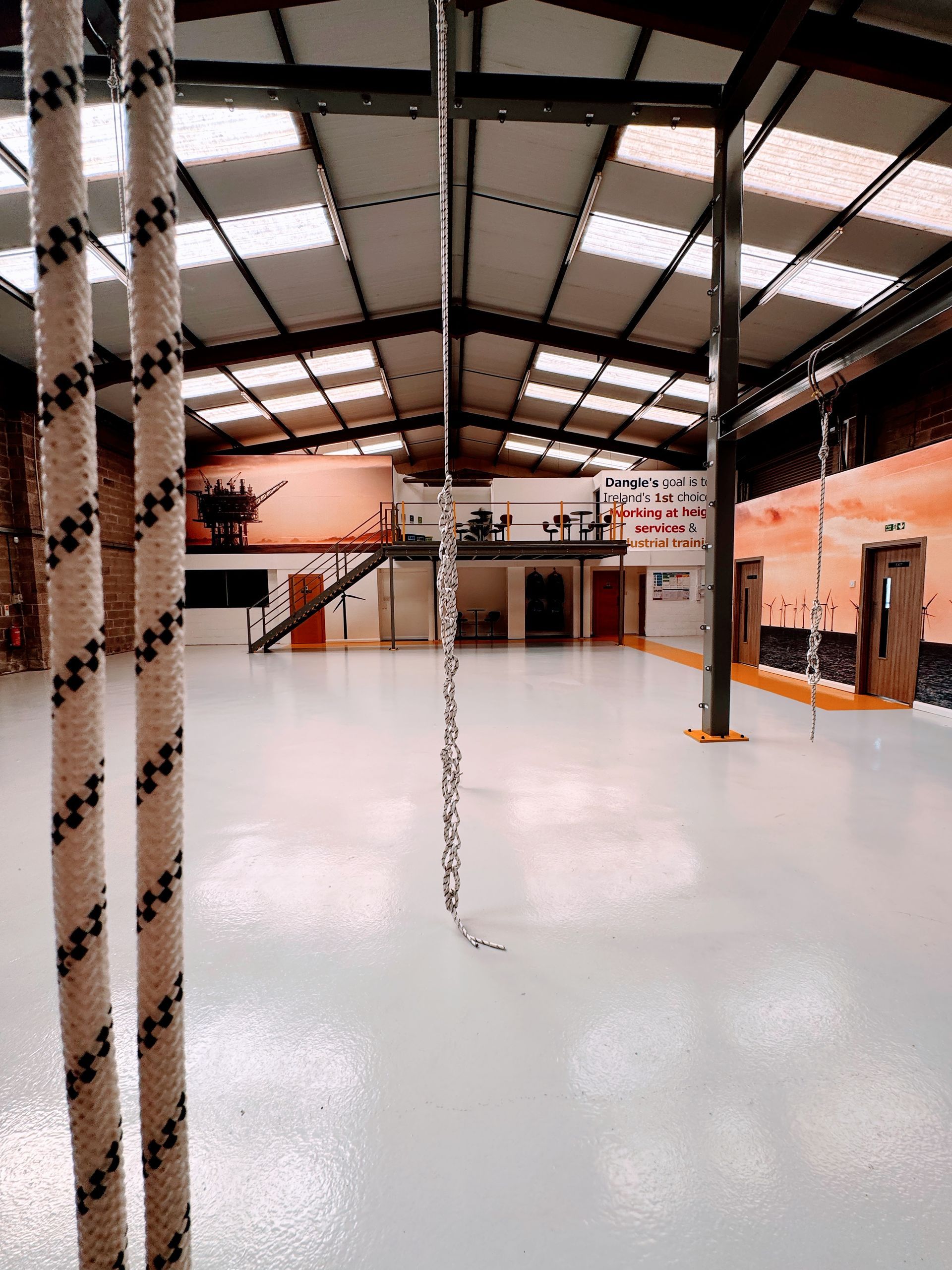 An open warehouse interior with a shiny white floor, ropes hanging from the high ceiling, and orange-painted walls.