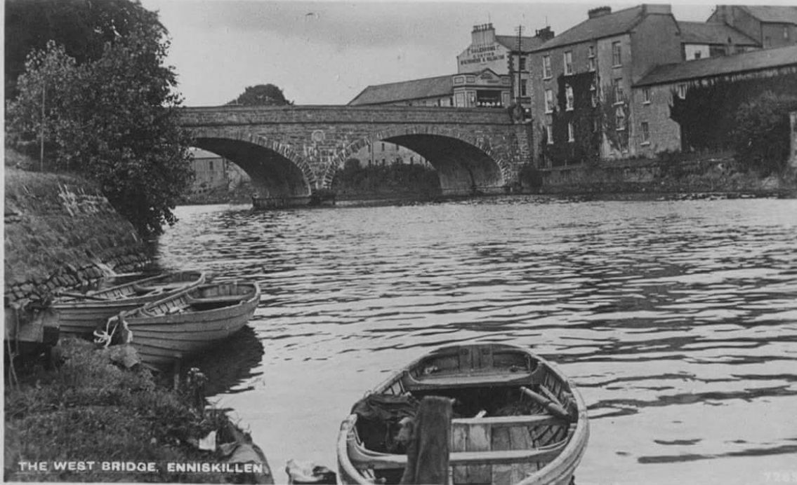 a old black and white photo of boats on a river under an arched stone bridge in Enniskillen.