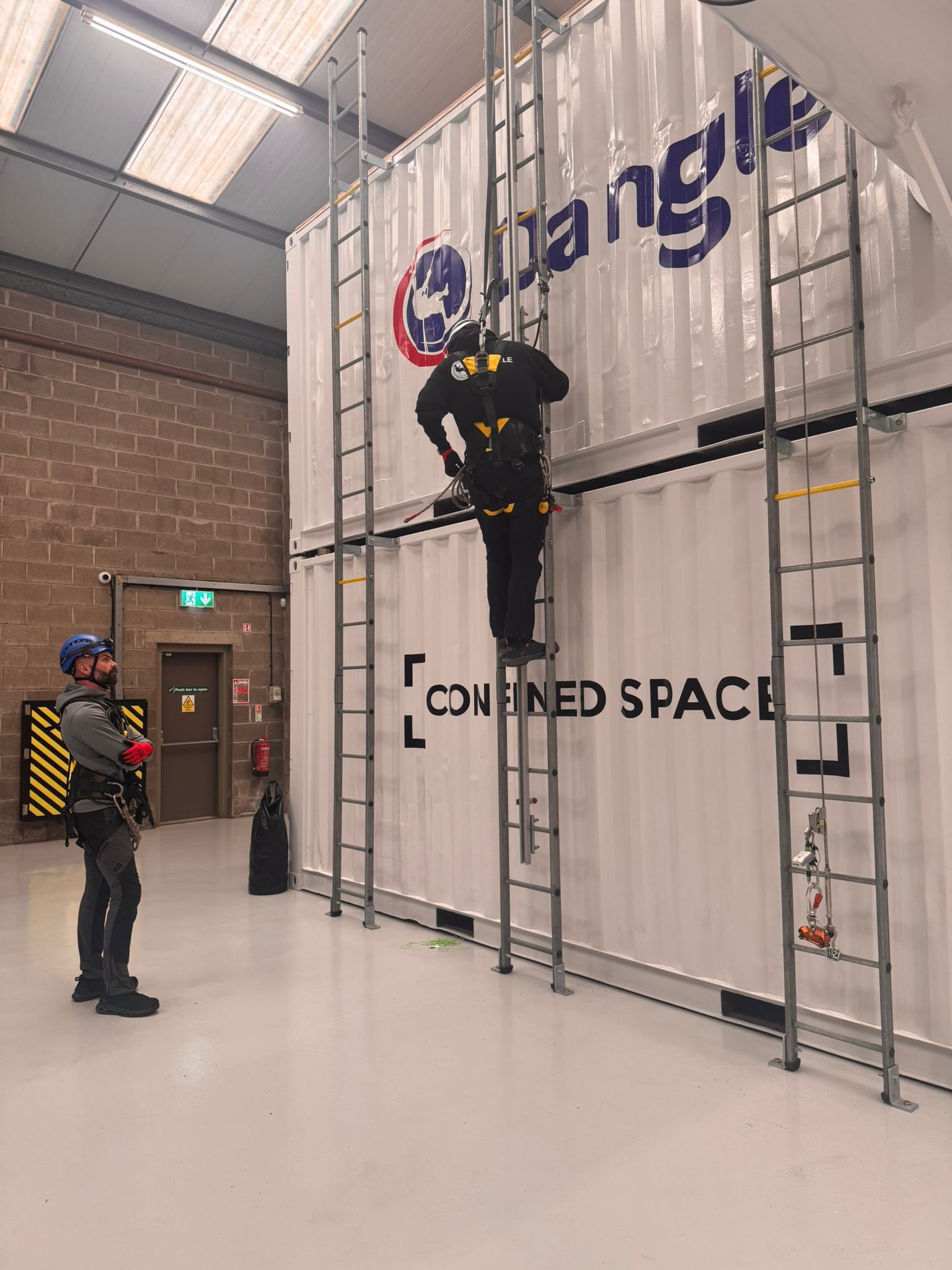 Worker climbing ladder in warehouse beside a large storage container labeled “CONFINED SPACE”