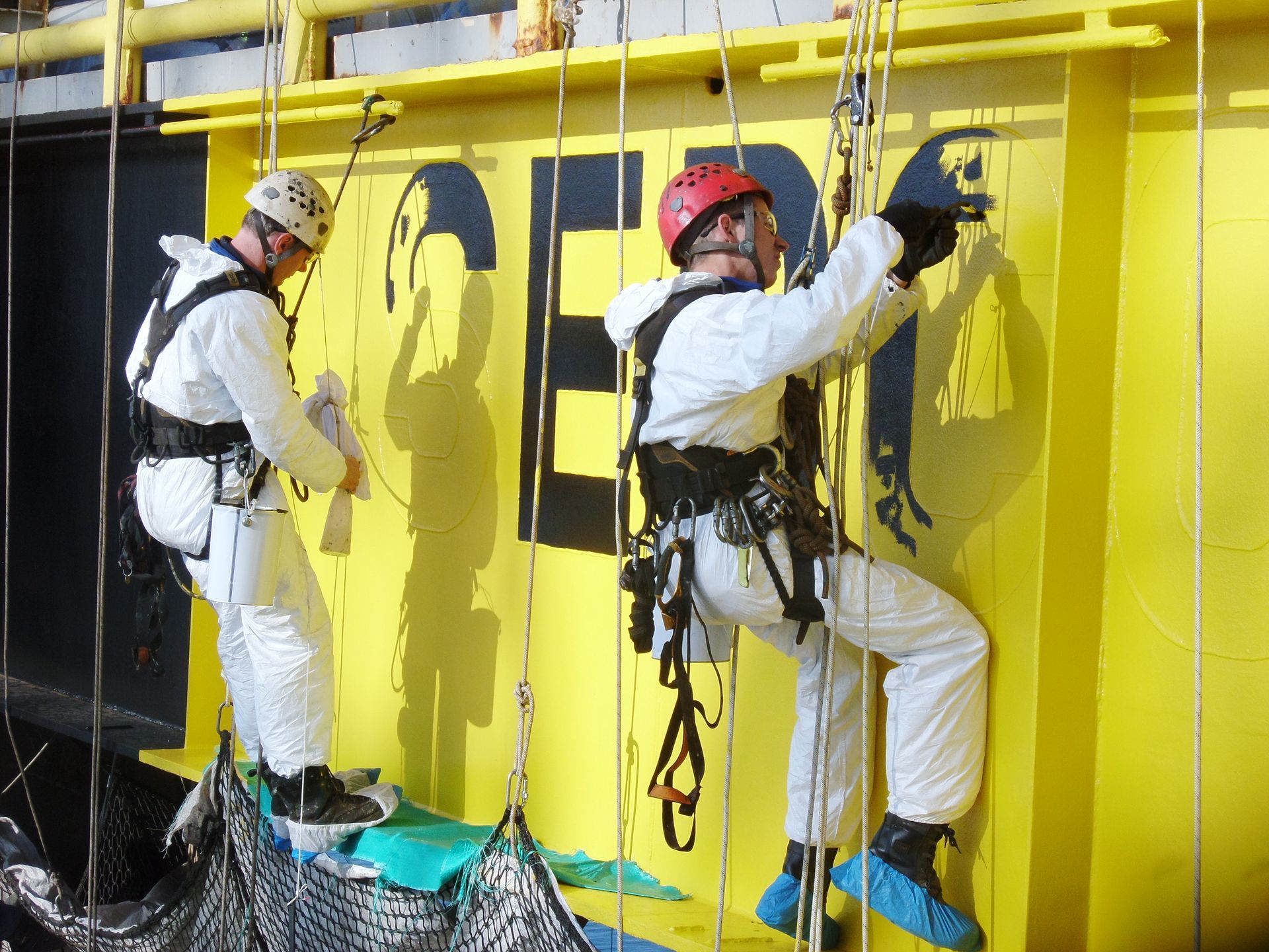 Two people in white protective suits hanging on ropes, painting a yellow wall.