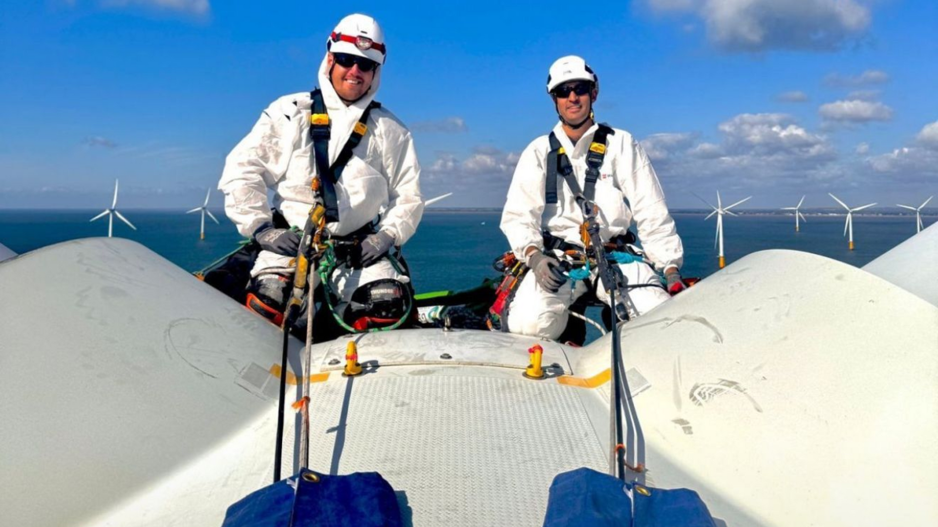 Two dangle rope-access workers on a wind turbine nacelle, wearing white suits and safety gear, overlooking ocean turbines.