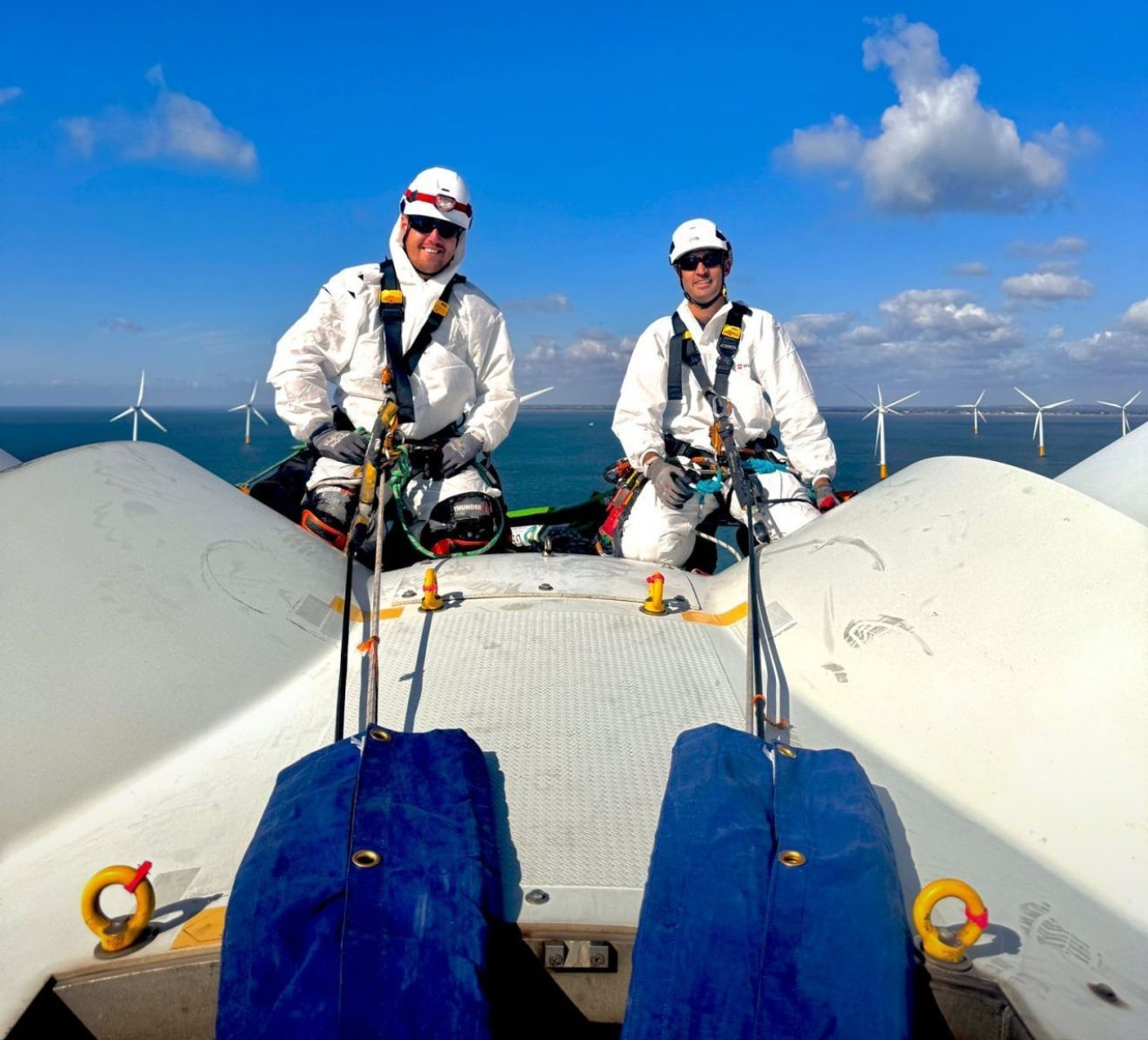 Two workers in white safety gear atop wind turbine blades against a blue sky starting out their rope access careers