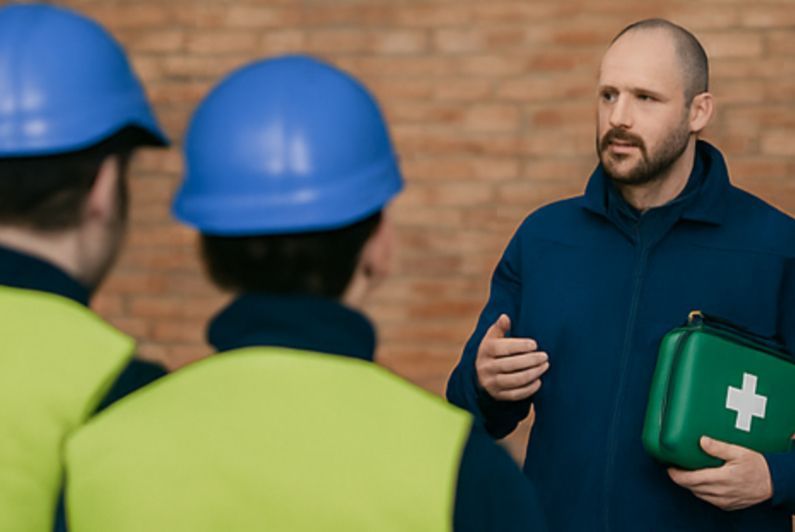 Man in blue uniform talking to two workers in hard hats and vests, holding a first aid kit.