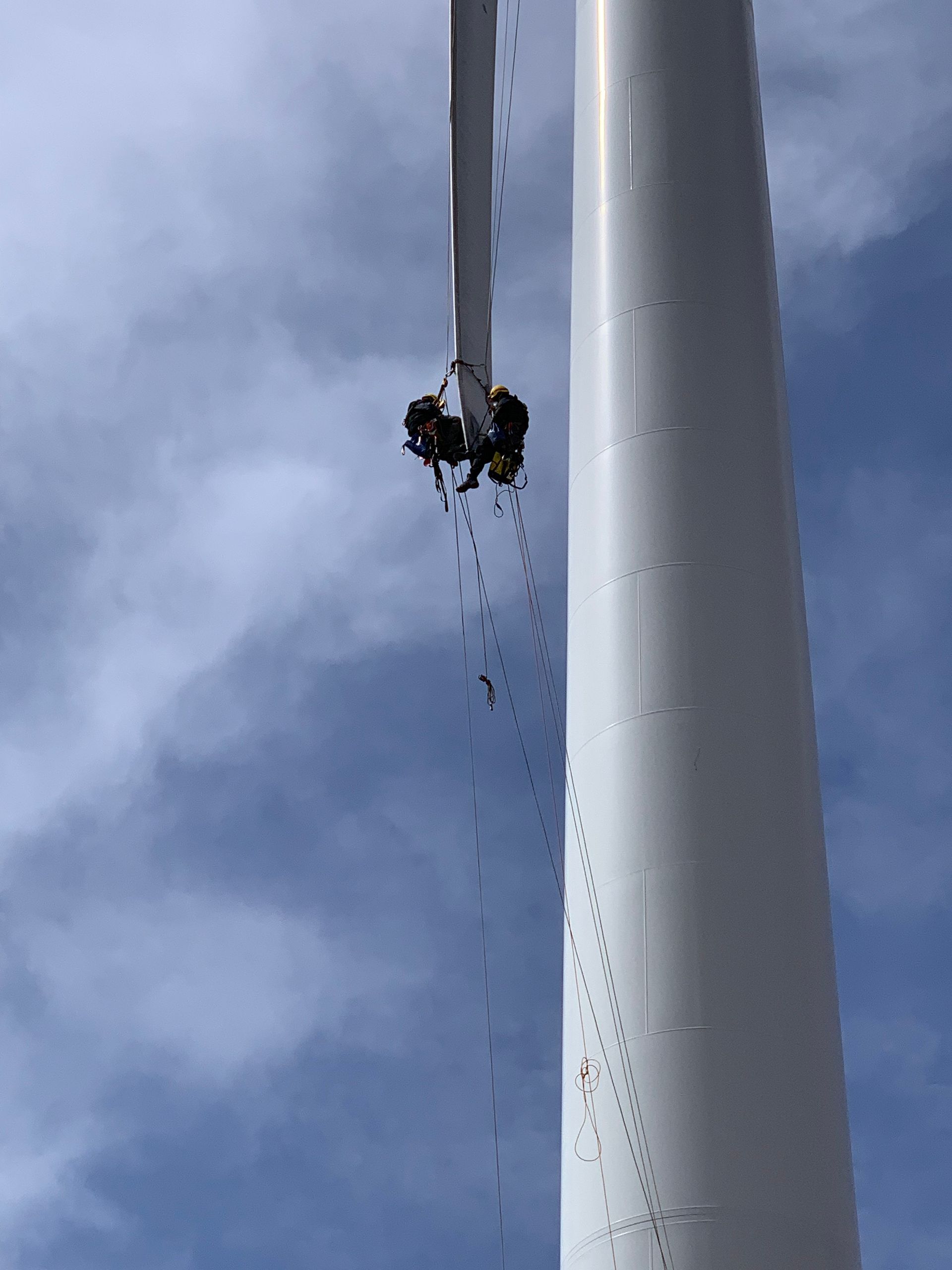 A group of people are using rope access to climb a white wind turbine.