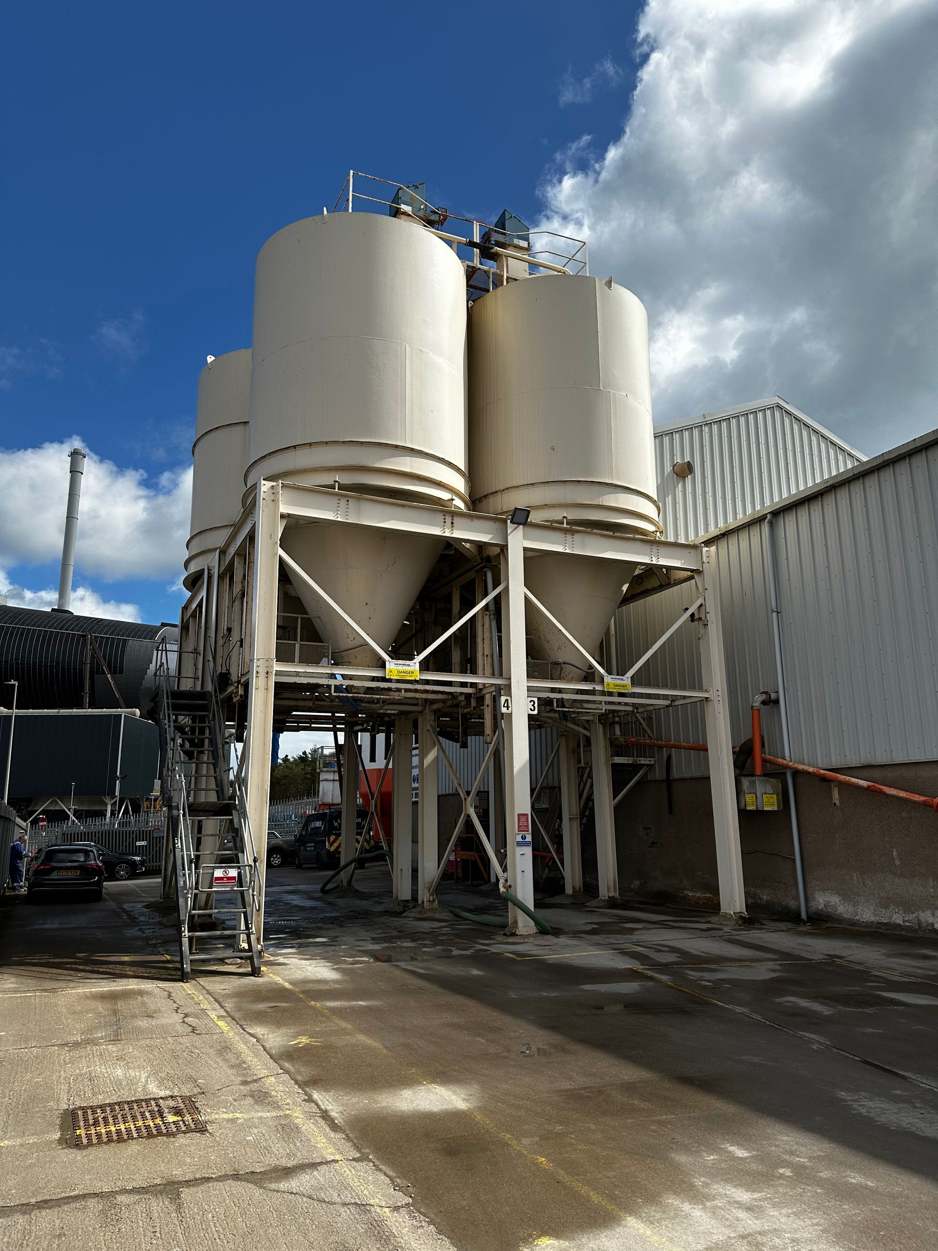 Three large white industrial silos on a metal support structure, next to a building, under a cloudy blue sky.