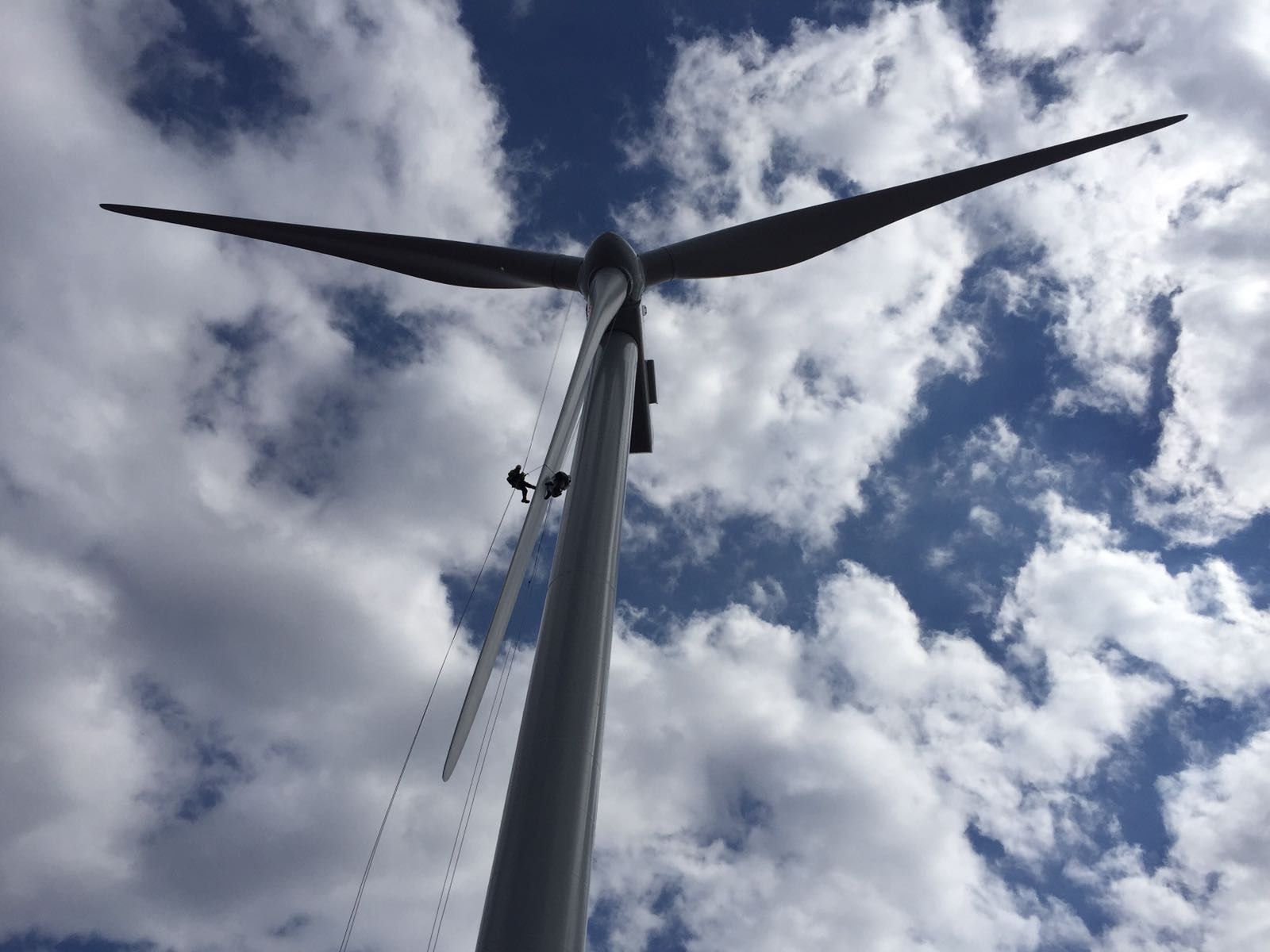 A wind turbine is against a cloudy blue sky with two technicians on the blade.