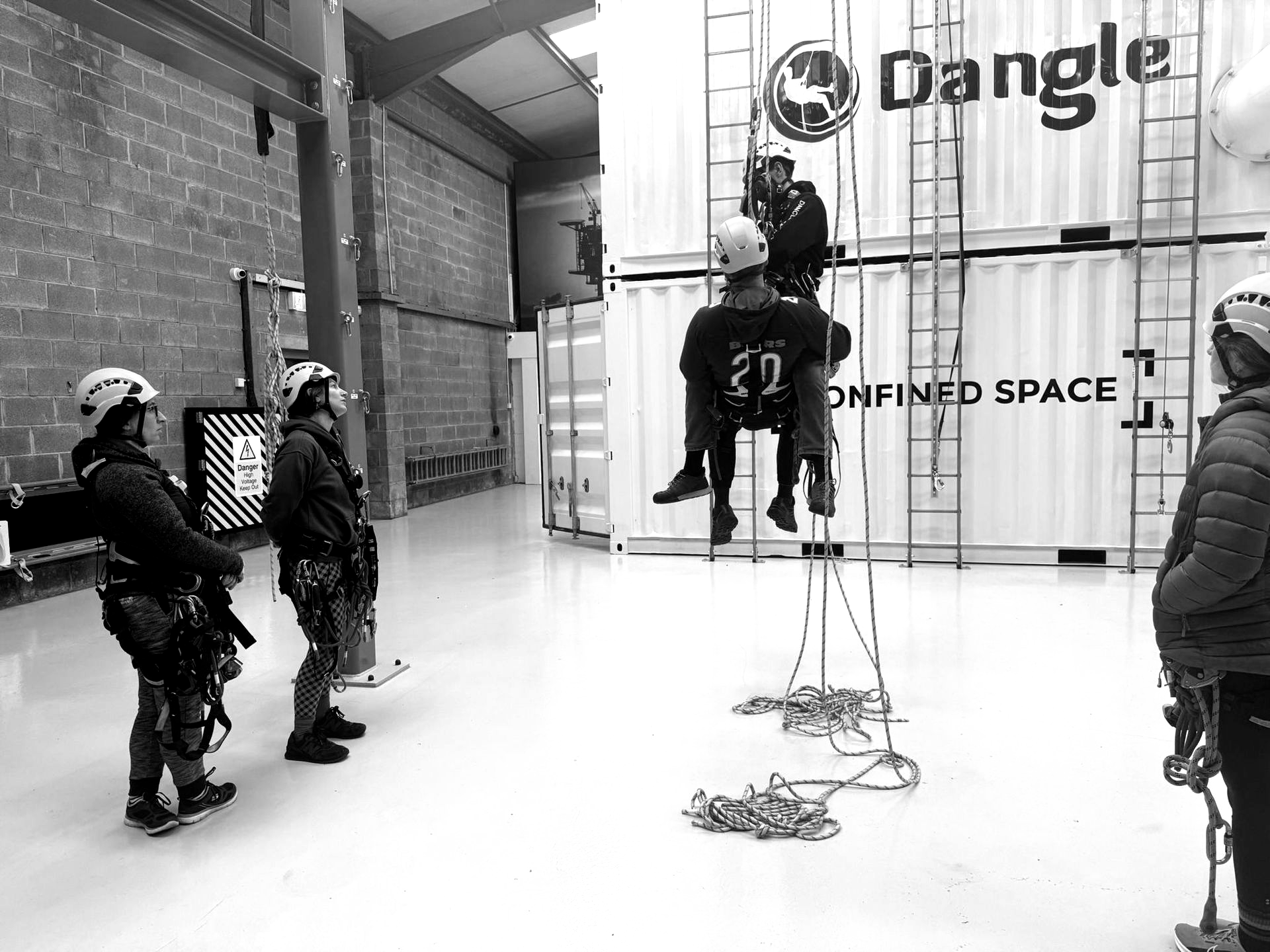 People in safety gear and helmets train on ropes in an industrial facility with shipping containers labeled