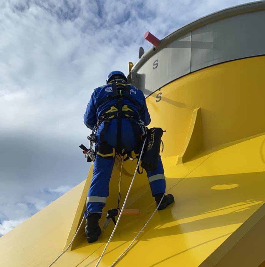 A man in a blue jumpsuit is standing on a yellow surface with a harness on.