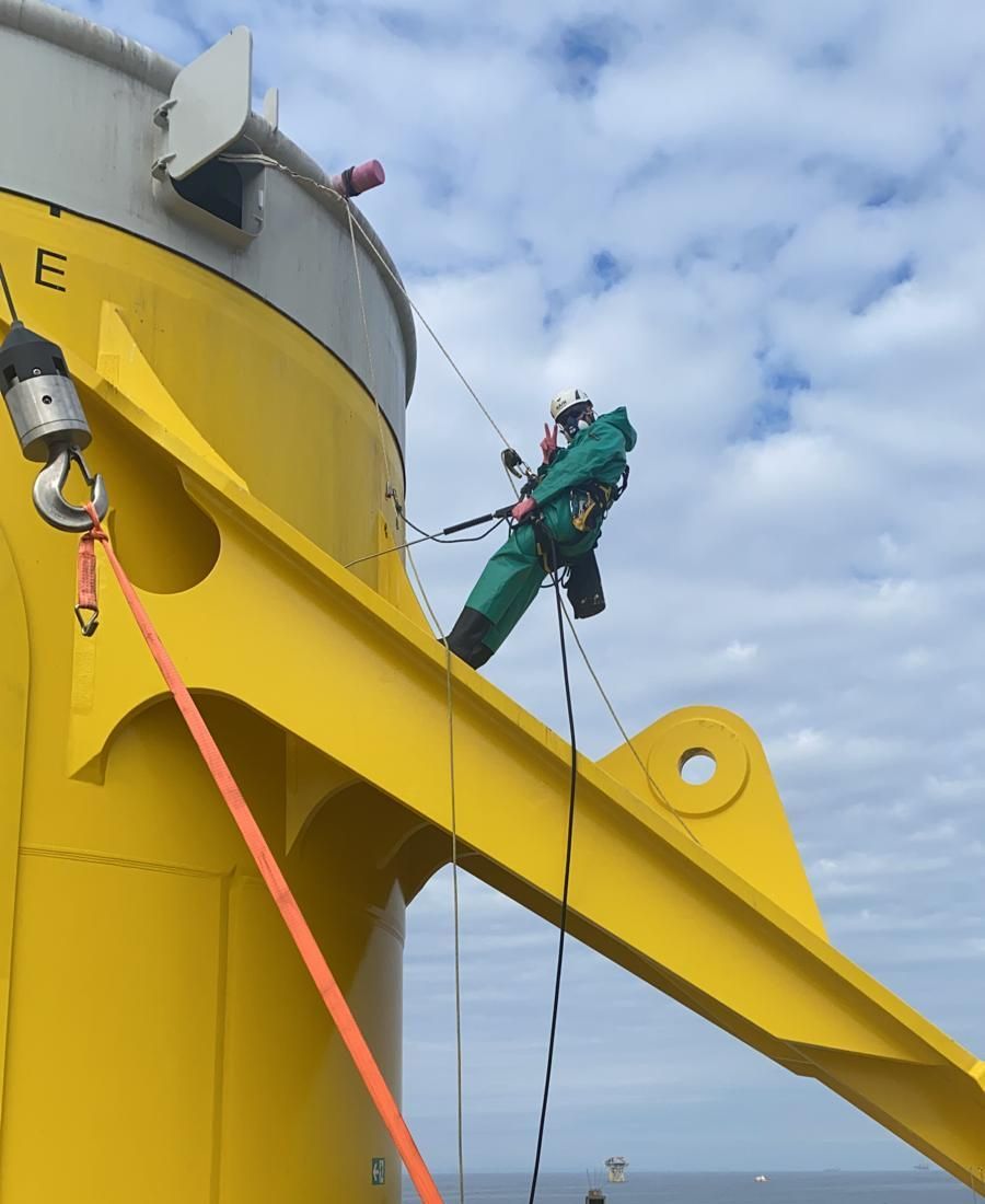Person in green protective suit rappelling down a yellow industrial structure, attached with ropes and harnesses.