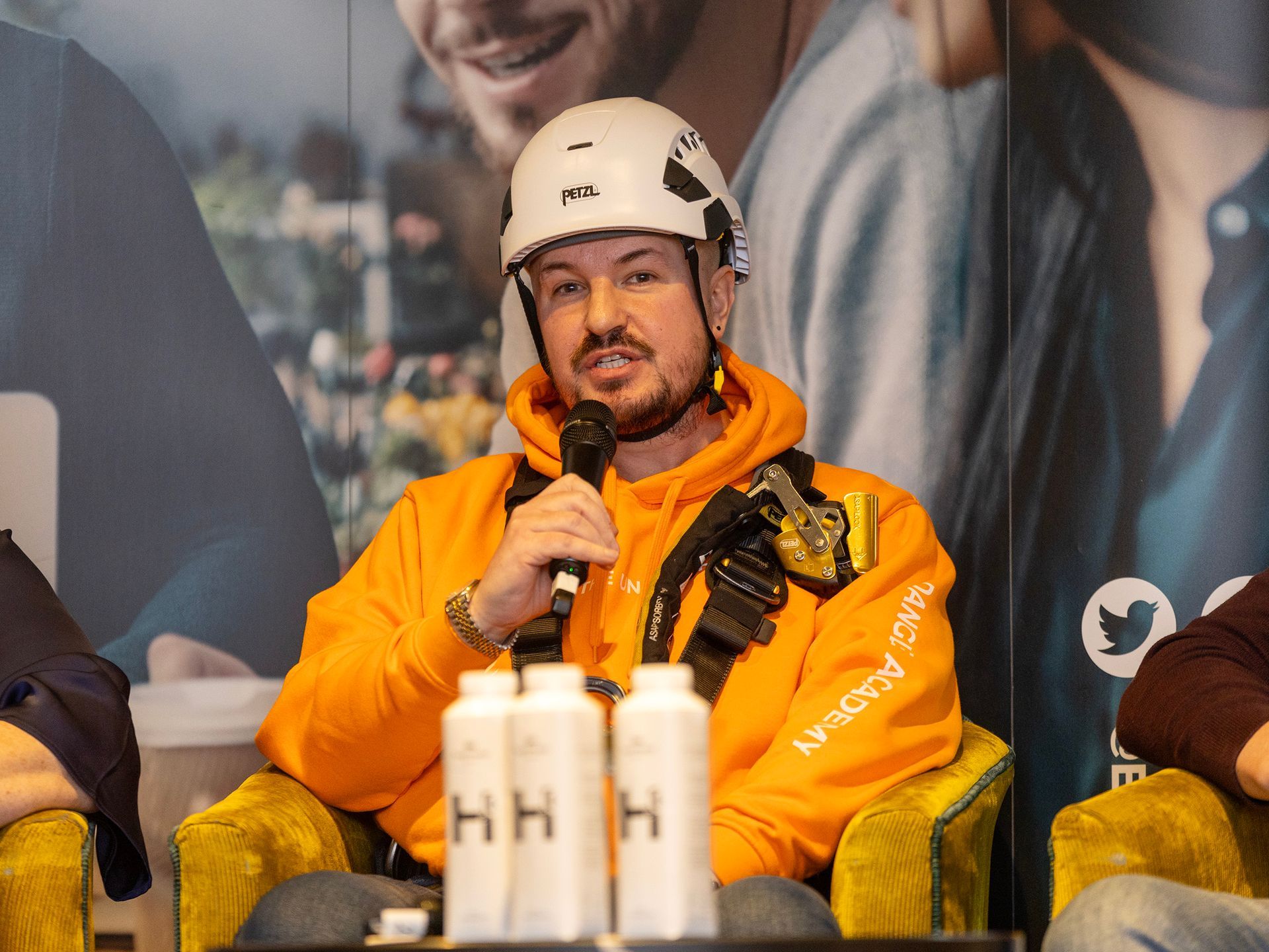 Simon in orange hoodie and white helmet speaking into a microphone. Seated, at a Enterprise NI event in Belfast.