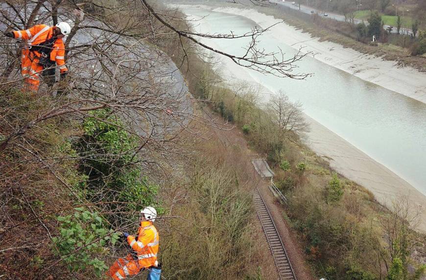 Two workers in orange safety gear trimming trees along a railway track beside a river.