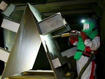 Person in protective gear sandblasting metal structure inside a dimly lit workshop.