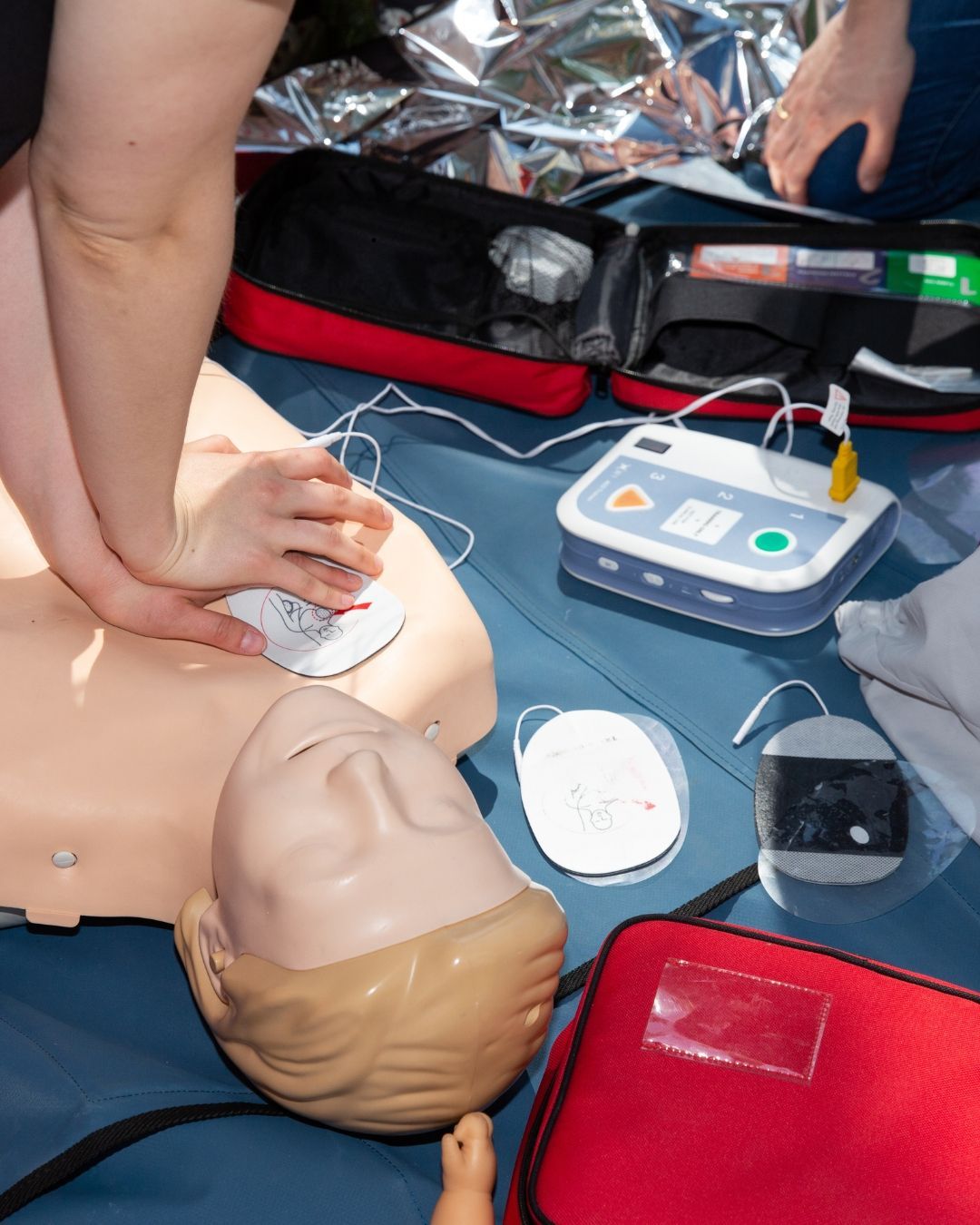 Hands performing CPR on a medical mannequin with an automated external defibrillator nearby.