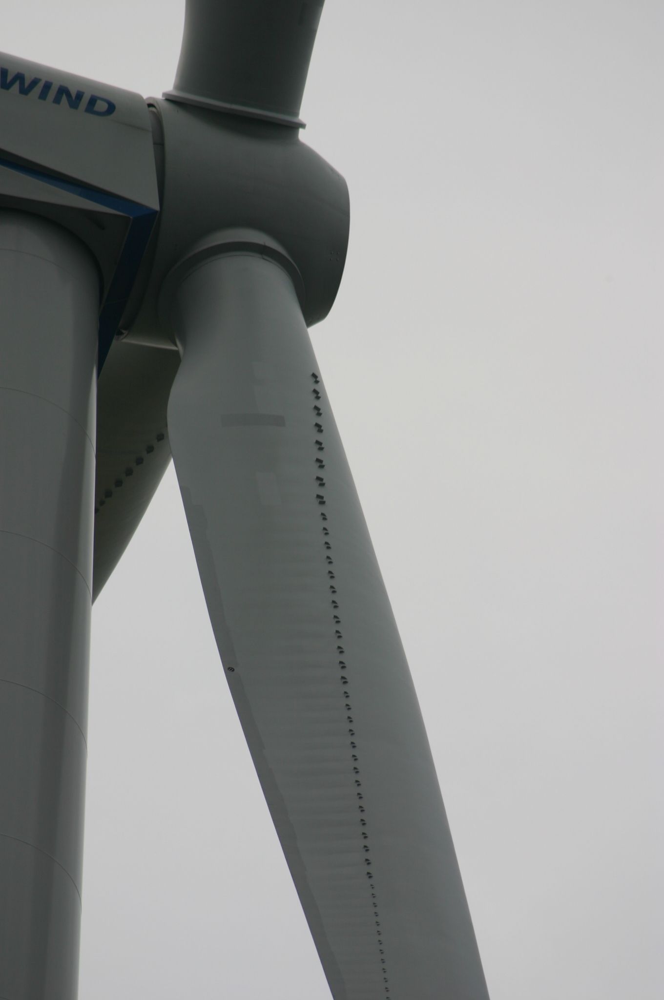 Close-up of a wind turbine blade with small openings along its side, against a cloudy sky.