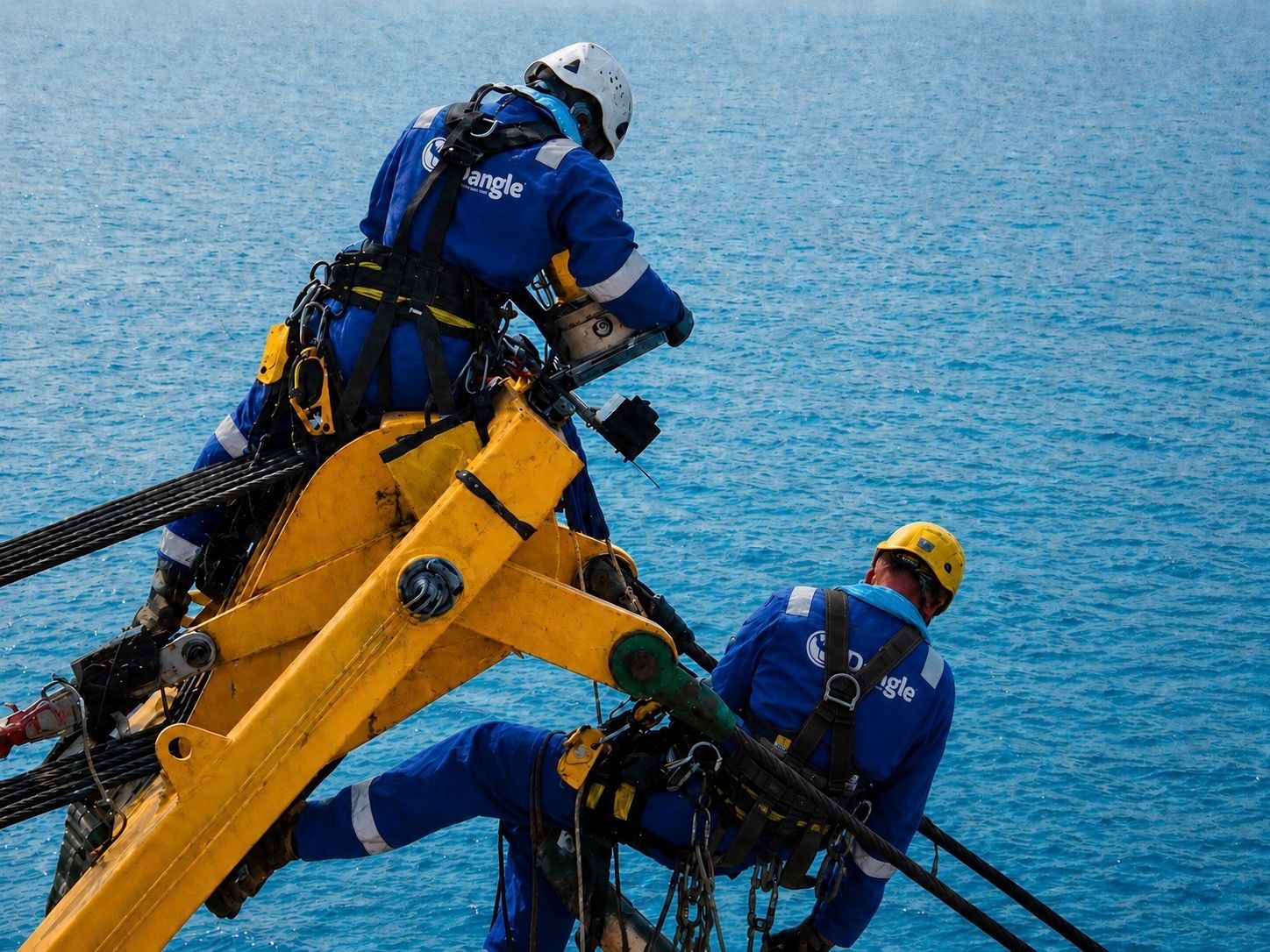 Two workers in blue gear on a yellow platform over the ocean, wearing safety harnesses and helmets.