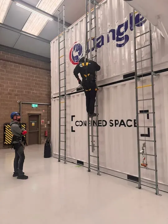 Worker climbing ladder in warehouse beside a large storage container labeled “CONFINED SPACE”