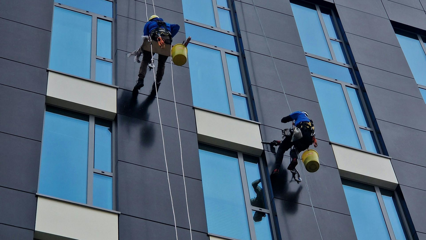 Two window washers on ropes cleaning windows of a dark coloured high-rise building in Belfast city centre.