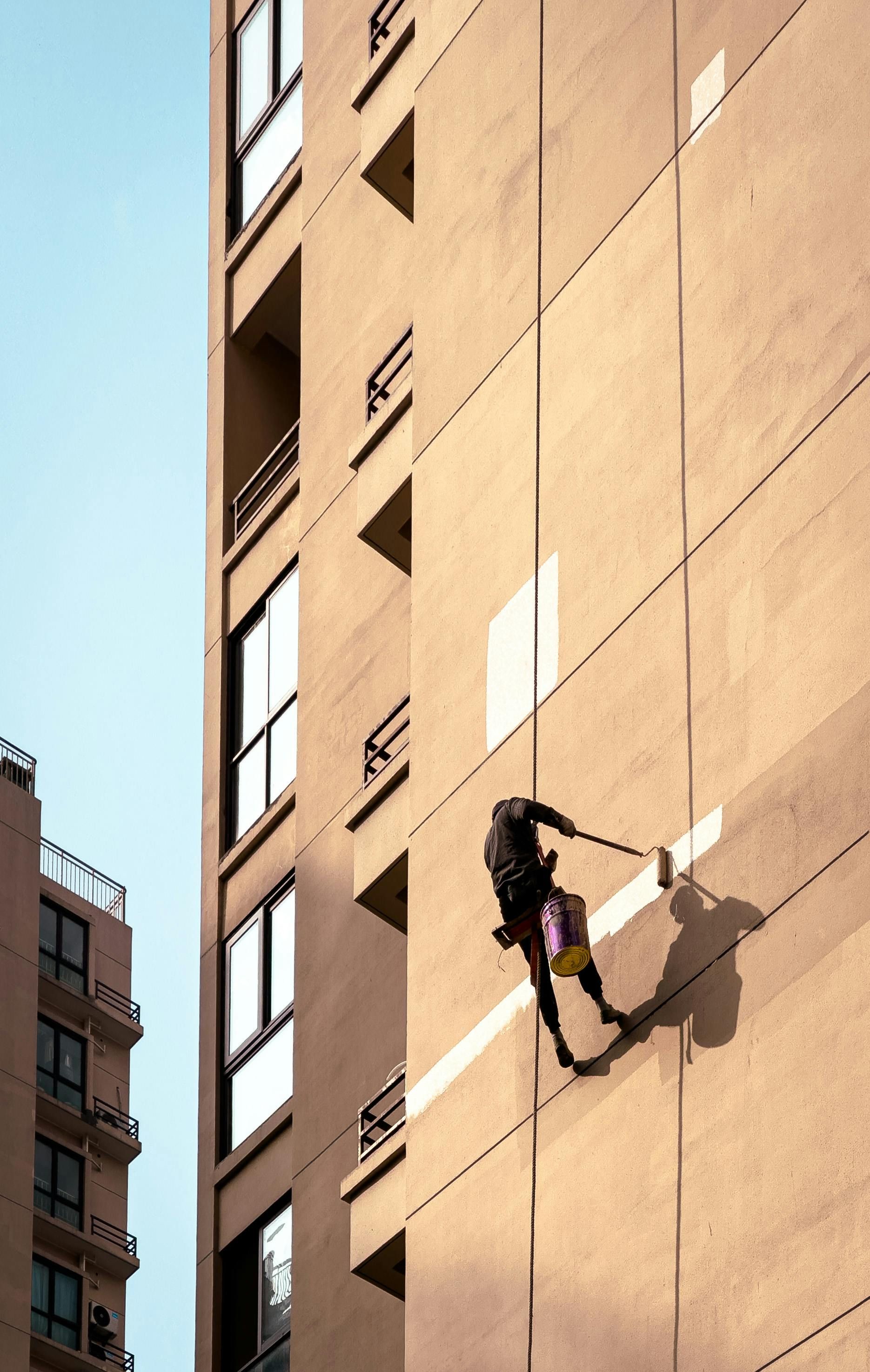 A person rappels down the side of a tall beige building, painting. They wear a harness and are attached to a rope.