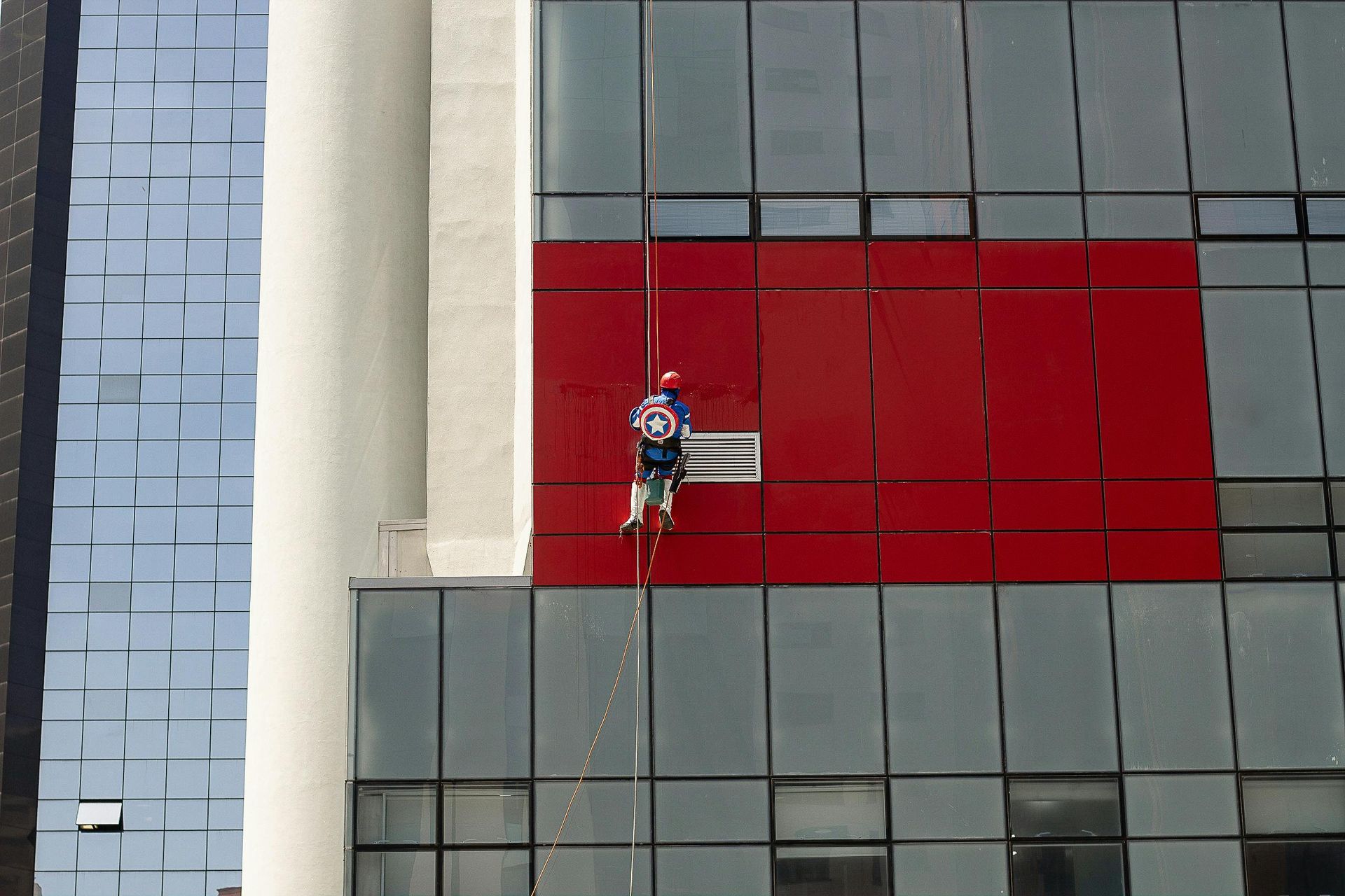 Window washer on a building's red and glass facade, secured by ropes, dressed as Captain America.