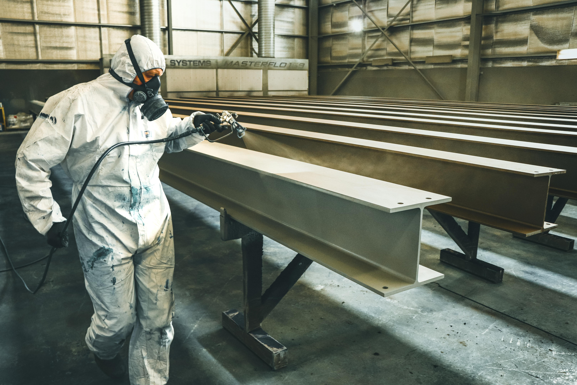 Person in protective suit spray applying intumescent paint on metal beams in a factory.