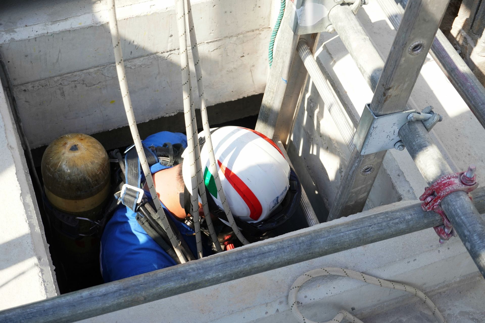 Person in blue suit and helmet descends a manhole, assisted by ropes and ladder.