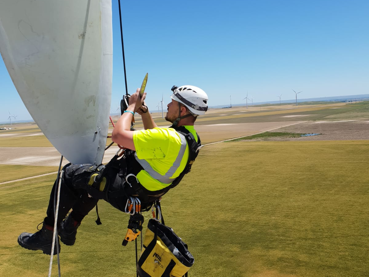 Person on wind turbine blade, wearing safety harness and helmet, inspecting equipment, bright green vest, sunny day.