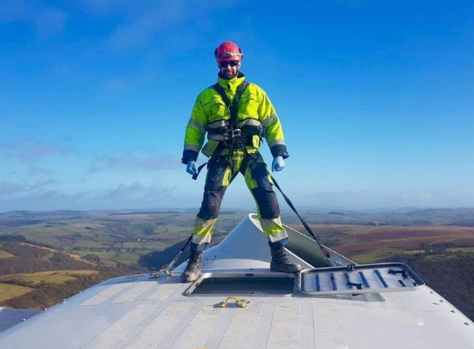 Safety-clad climber stands on a tall structure above a wide landscape, holding ropes under a blue sky.