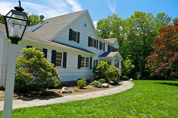 White two-story house with black shutters, green lawn, trees, and a pathway under a blue sky. White two-story house with black shutters, green lawn, trees, and a pathway under a blue sky.