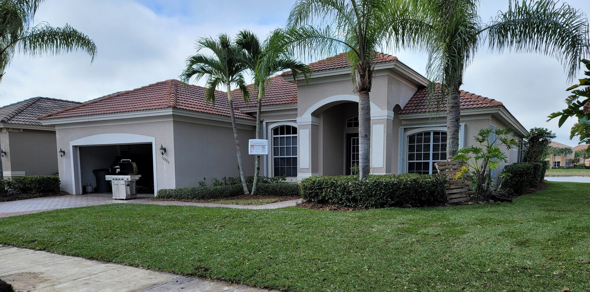 A beige house with a red-tiled roof, palm trees, and green grass. Cloudy sky.