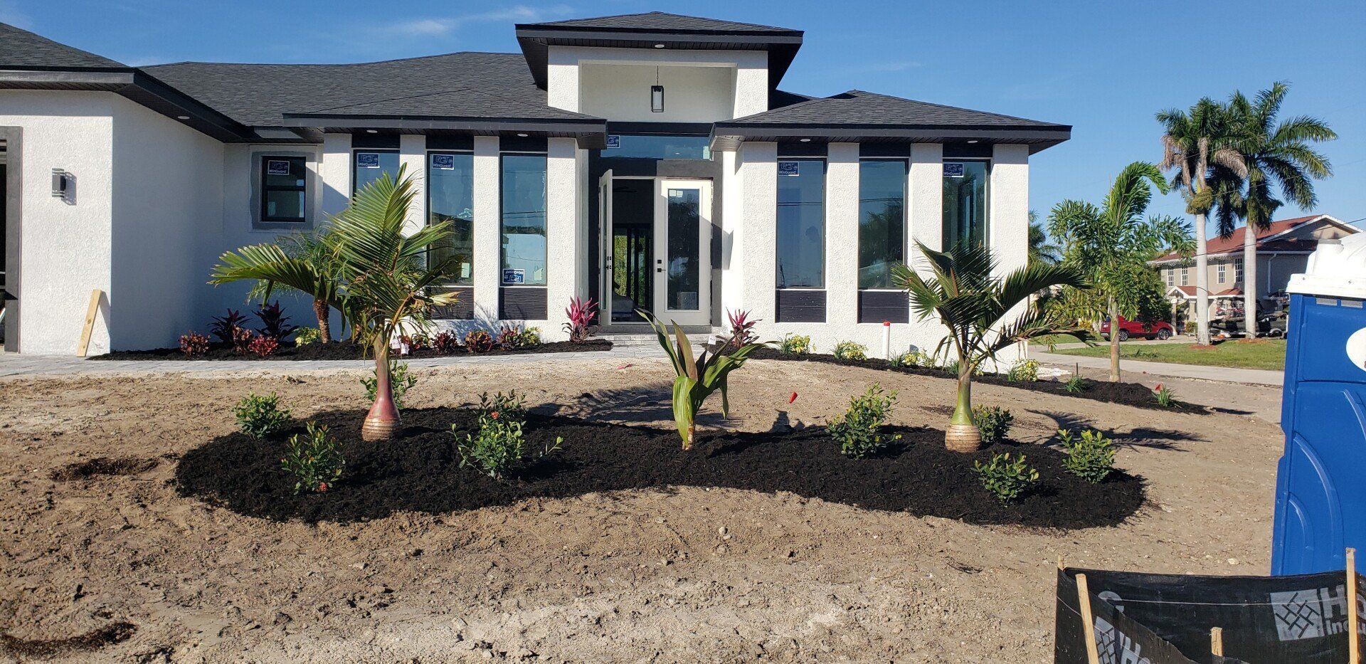 A modern white house with black roof, small palms, and landscaped bed on a sunny day.