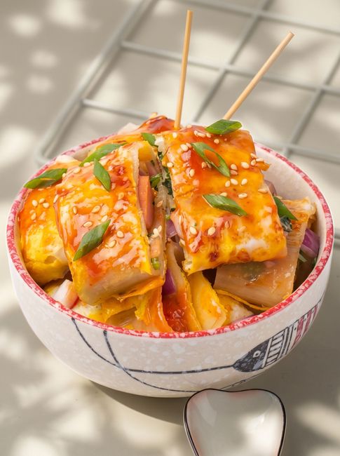Bowl of saucy tofu cubes topped with sesame seeds and herbs, with toothpicks on a checkered tablecloth