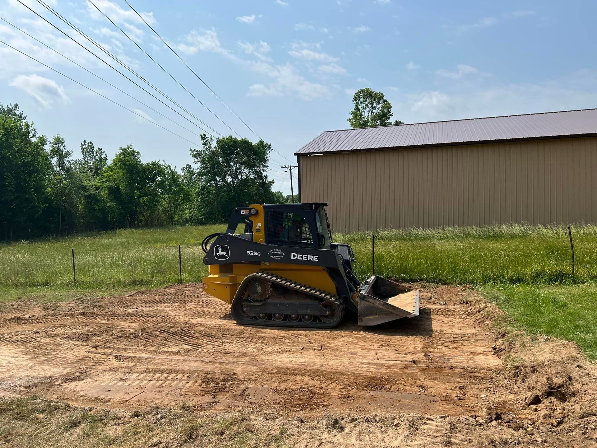 Yellow bulldozer grading a dirt lot beside a barn under a blue sky