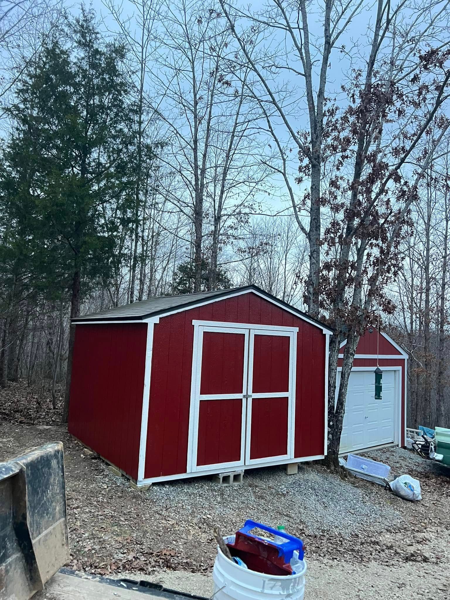 Red shed and white storage shed in a wooded backyard, with a bin in the foreground.