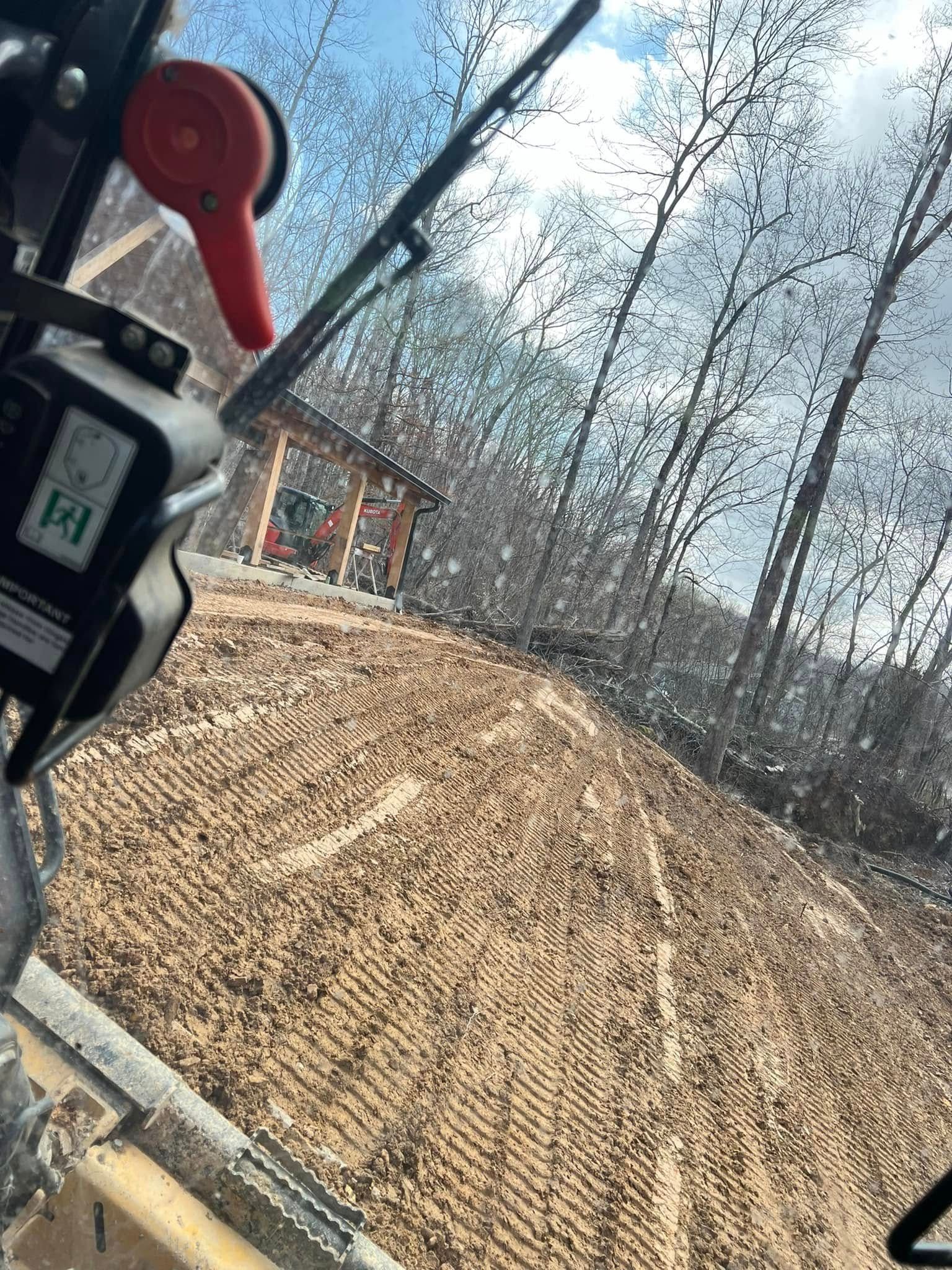 Construction site with freshly graded dirt and trees under a partly cloudy sky, viewed from inside a vehicle.