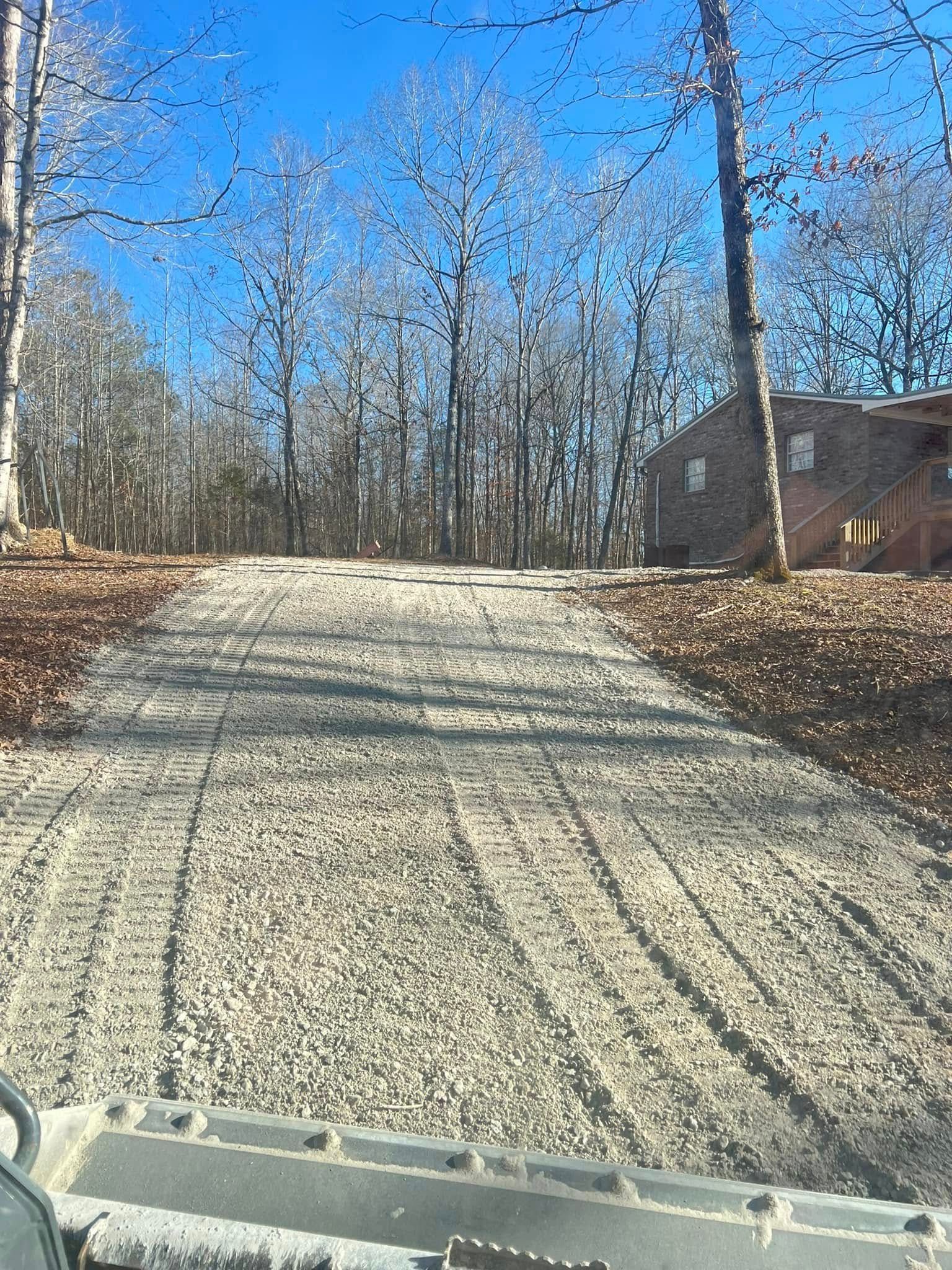 Gravel driveway on a sunny wooded hillside, with a cabin on the right and clear blue sky.