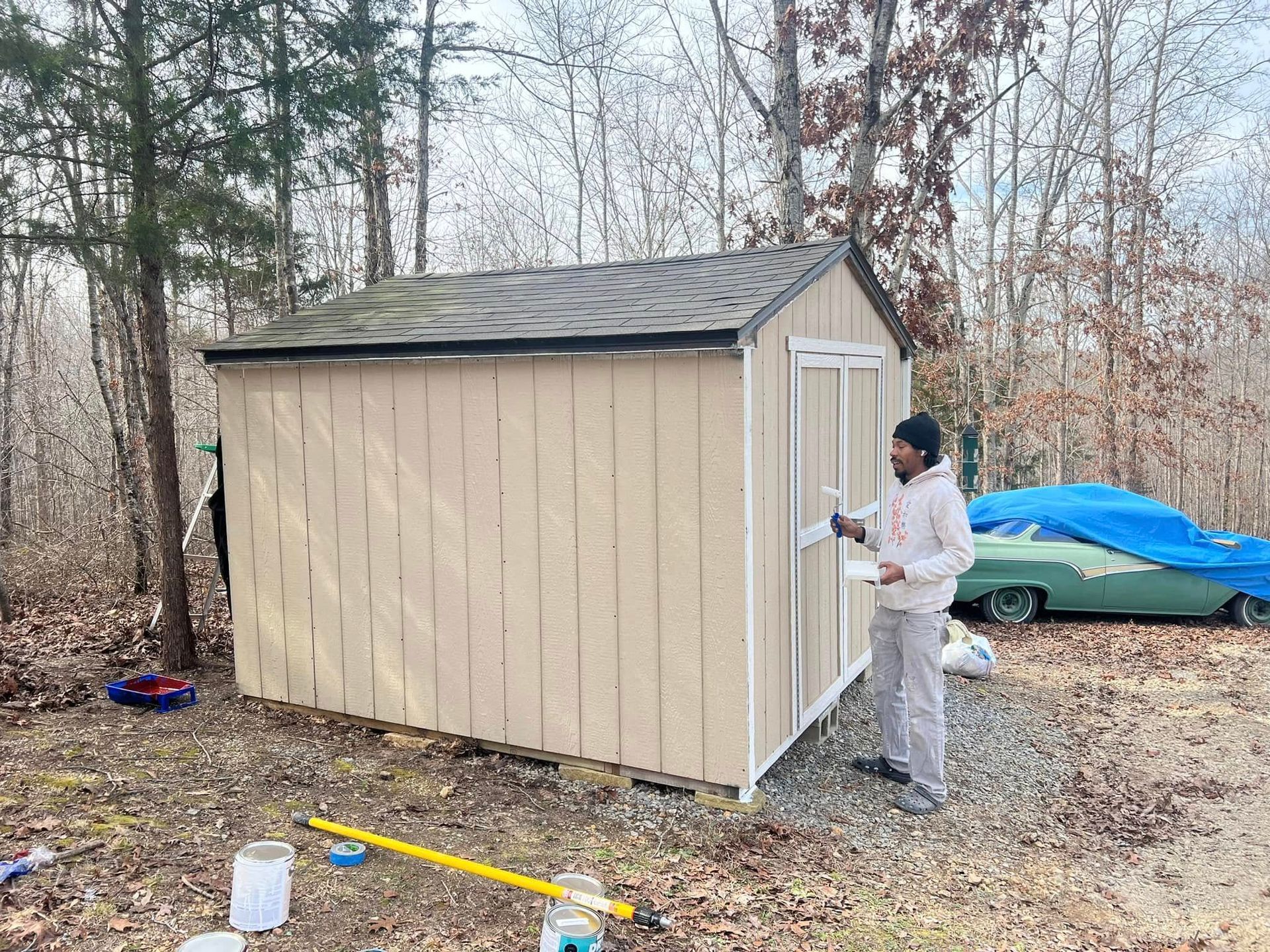 Person painting a beige shed in a wooded yard, with supplies and a covered boat nearby.