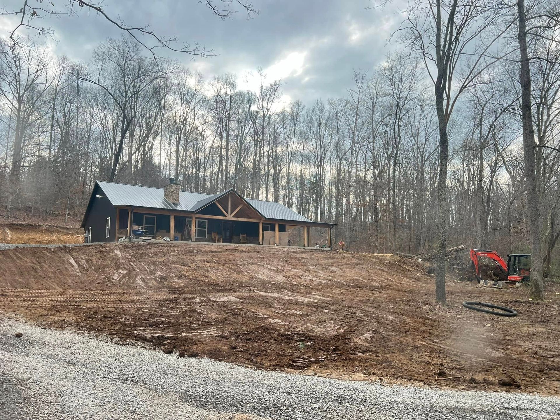 Rural house under construction in a wooded clearing, with bare trees and a red excavator on the right.