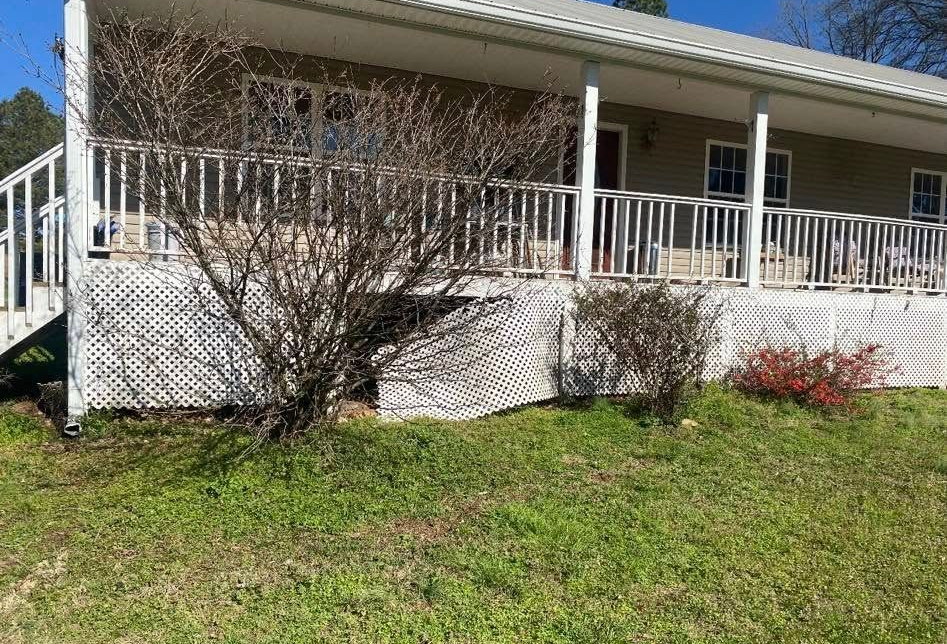 Front porch of a beige house with white railing, bushes, and a grassy yard under blue sky