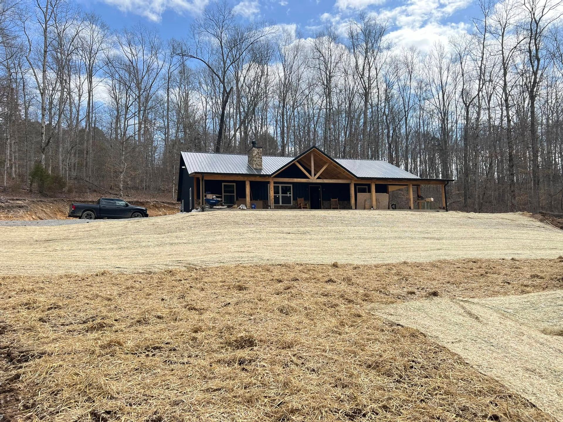 Rustic cabin with a metal roof in a clearing, surrounded by leafless trees under a cloudy blue sky
