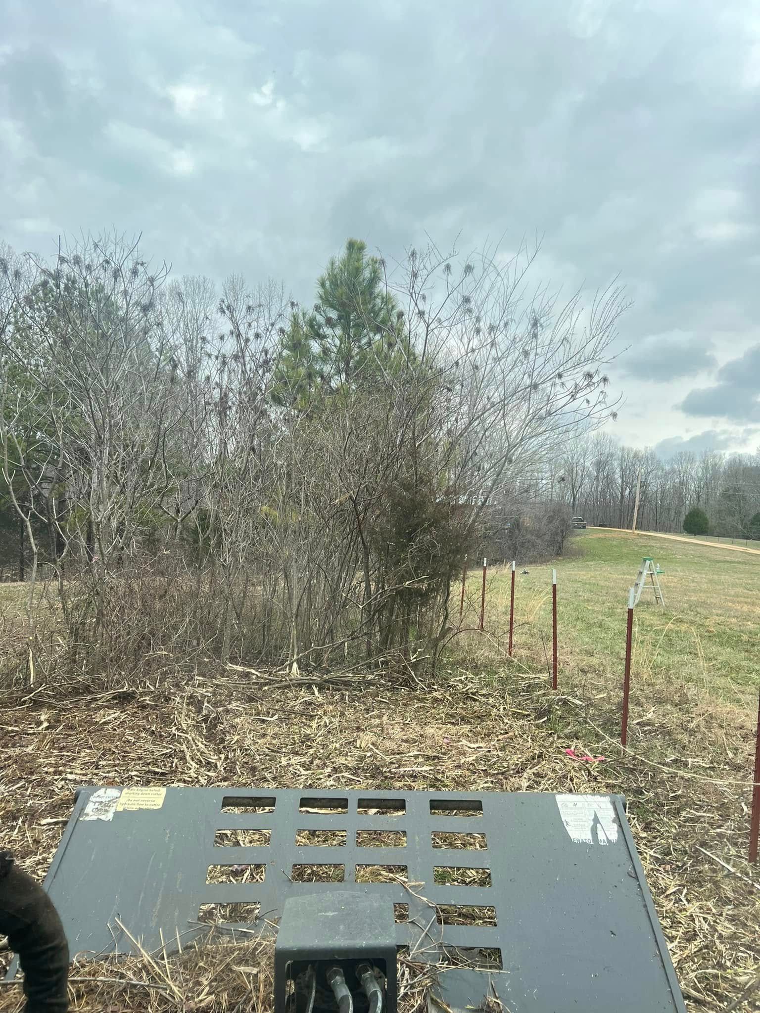 View from a tractor over a field with brush, fence posts, and a cloudy sky