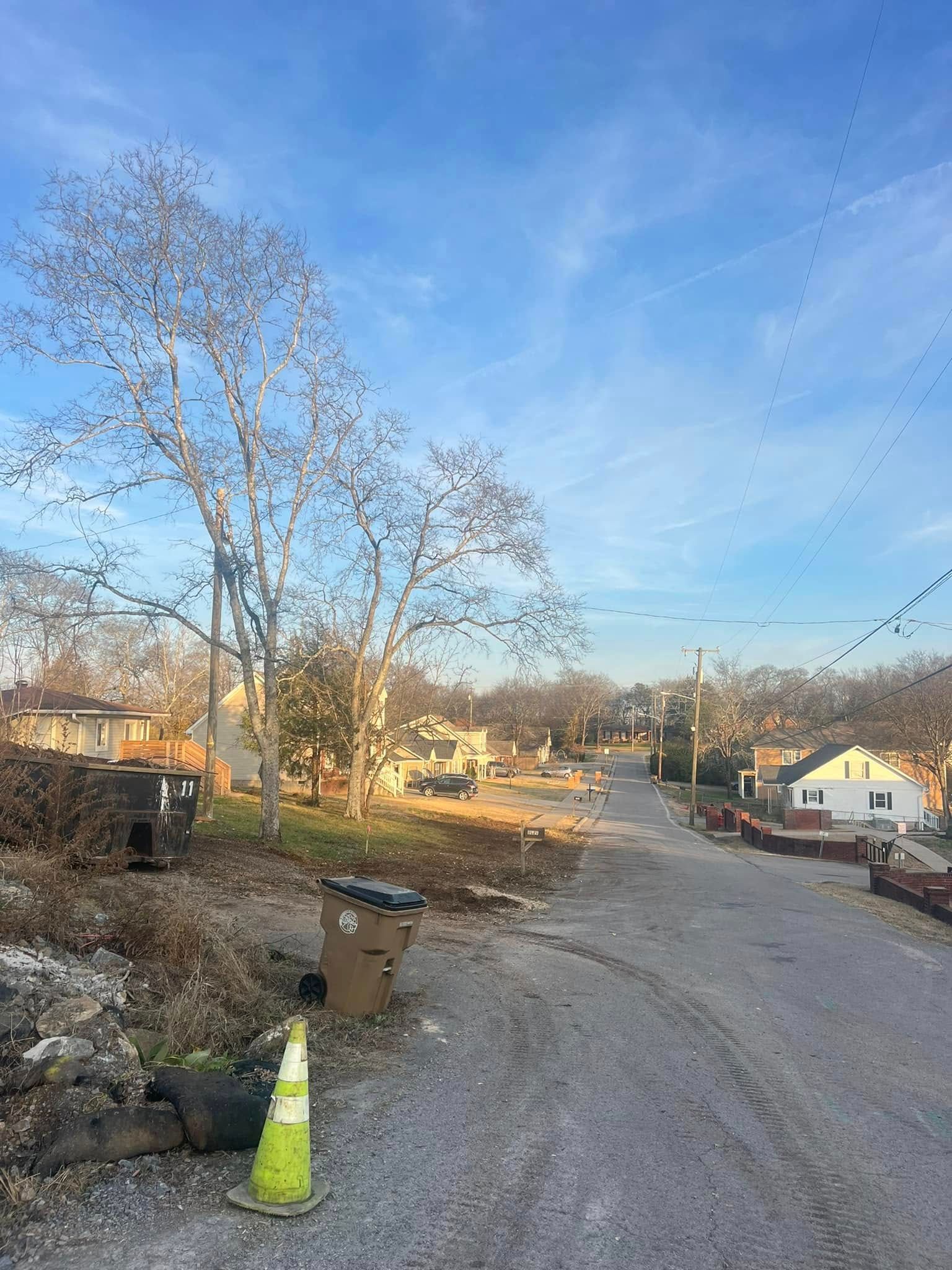 Rural gravel road with a traffic cone, leafless trees, and houses under a blue sky