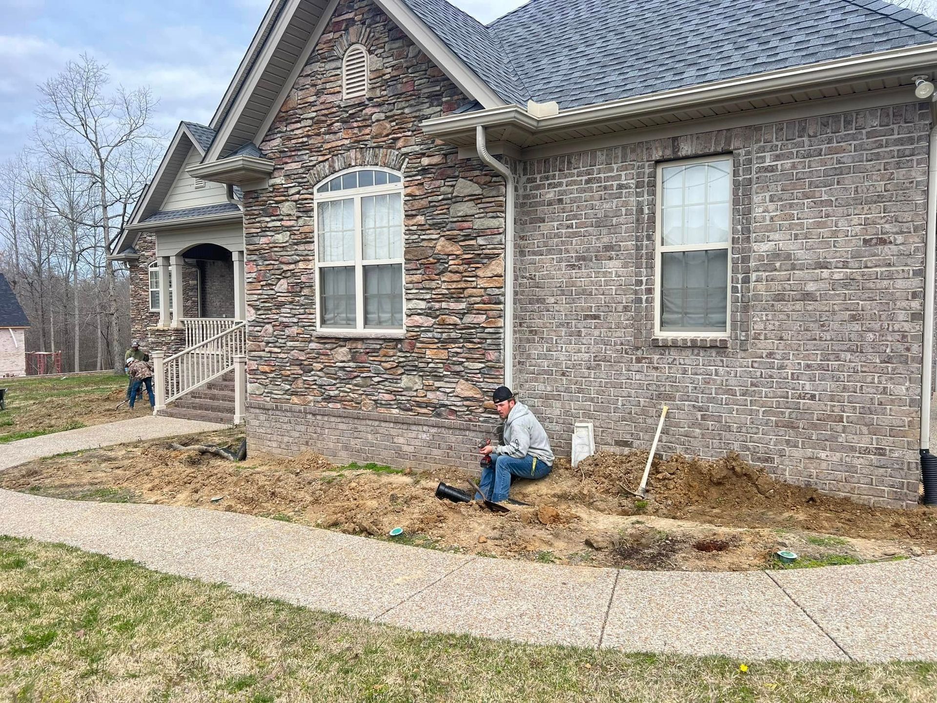 Worker kneeling by a brick house exterior, tending a mulched front planting bed near a sidewalk.